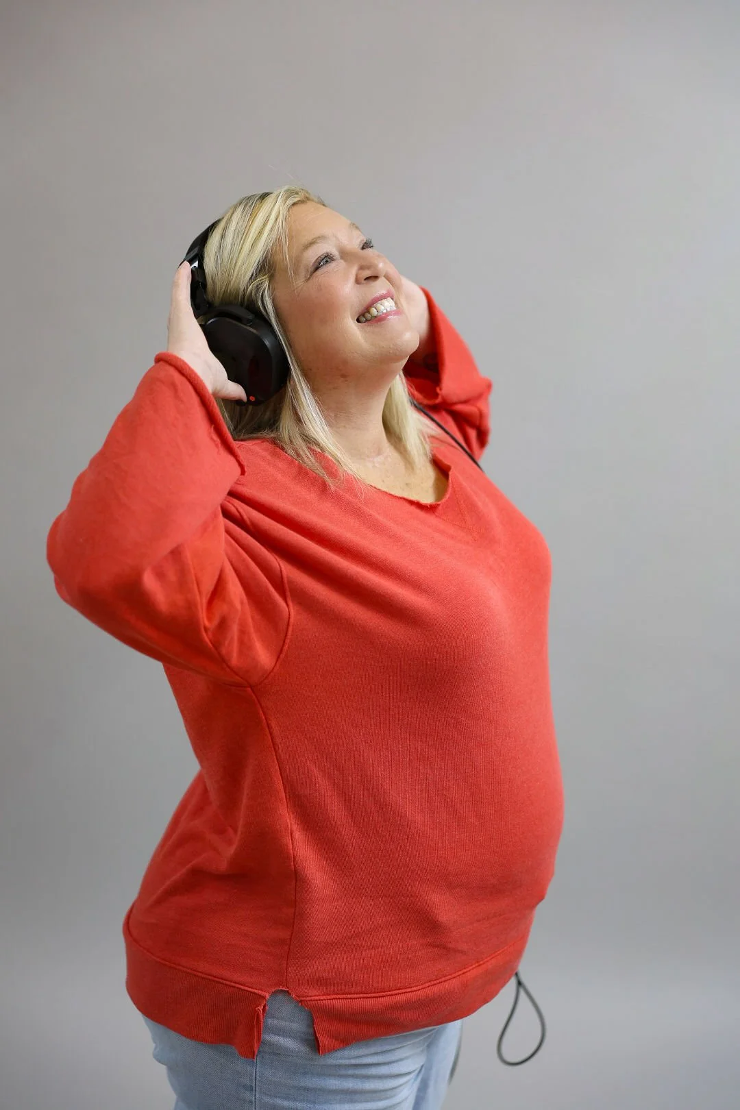 A woman with blonde hair wearing a red sweatshirt and jeans, smiling with eyes closed, holding large black headphones on her head against a plain gray wall.