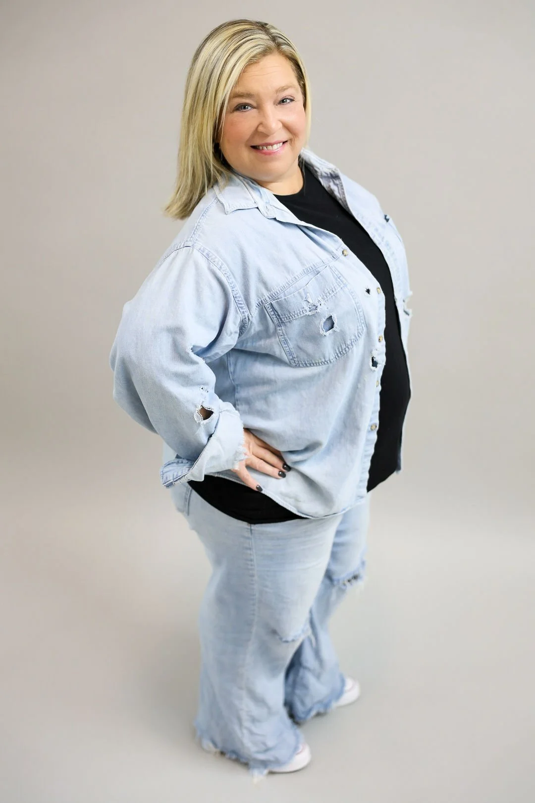 Woman in distressed denim jacket and jeans, smiling, standing with one hand on her hip against a neutral background.