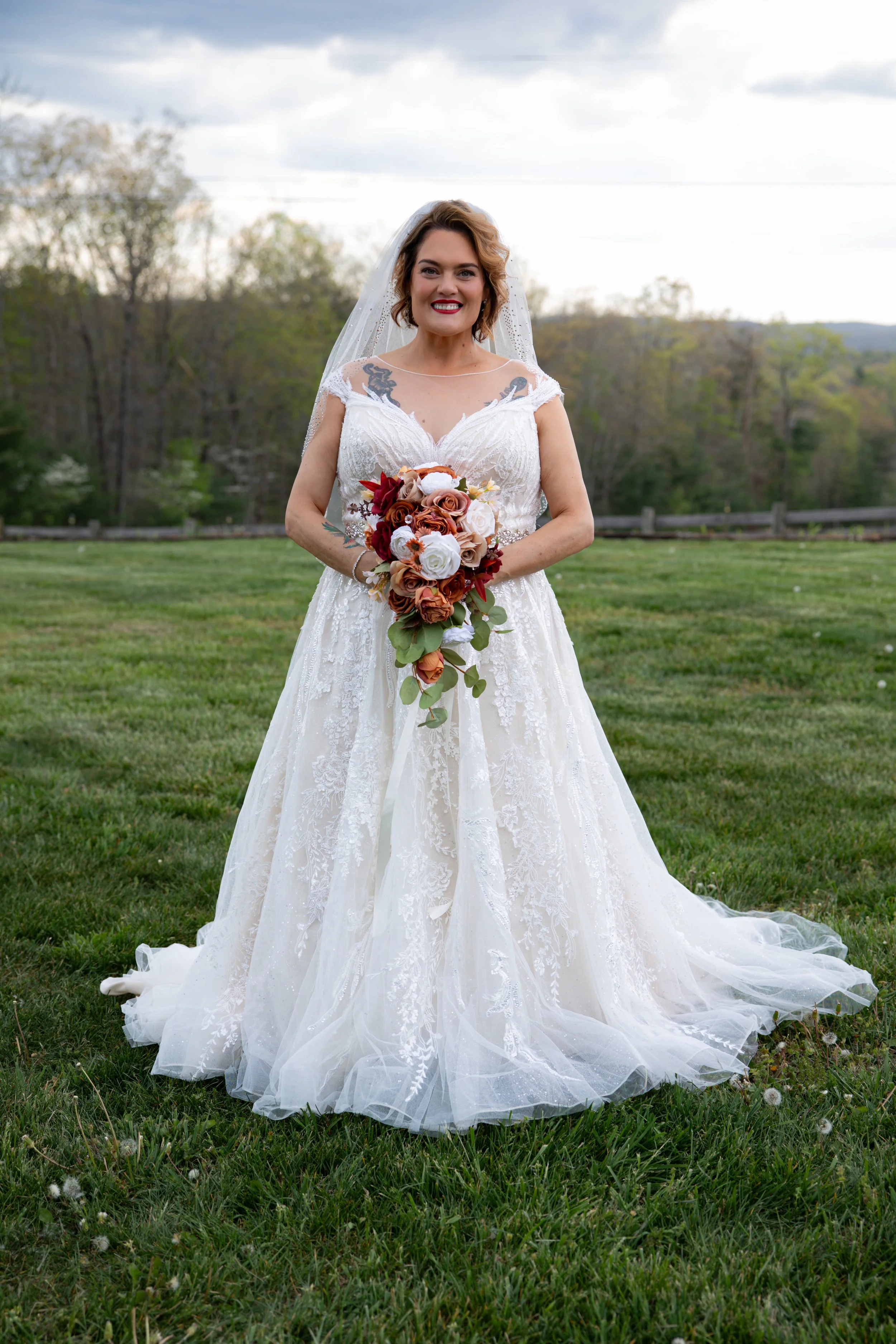 A bride in a white lace wedding dress holding a bouquet of roses and greenery, standing outdoors on grass with trees and a cloudy sky in the background.