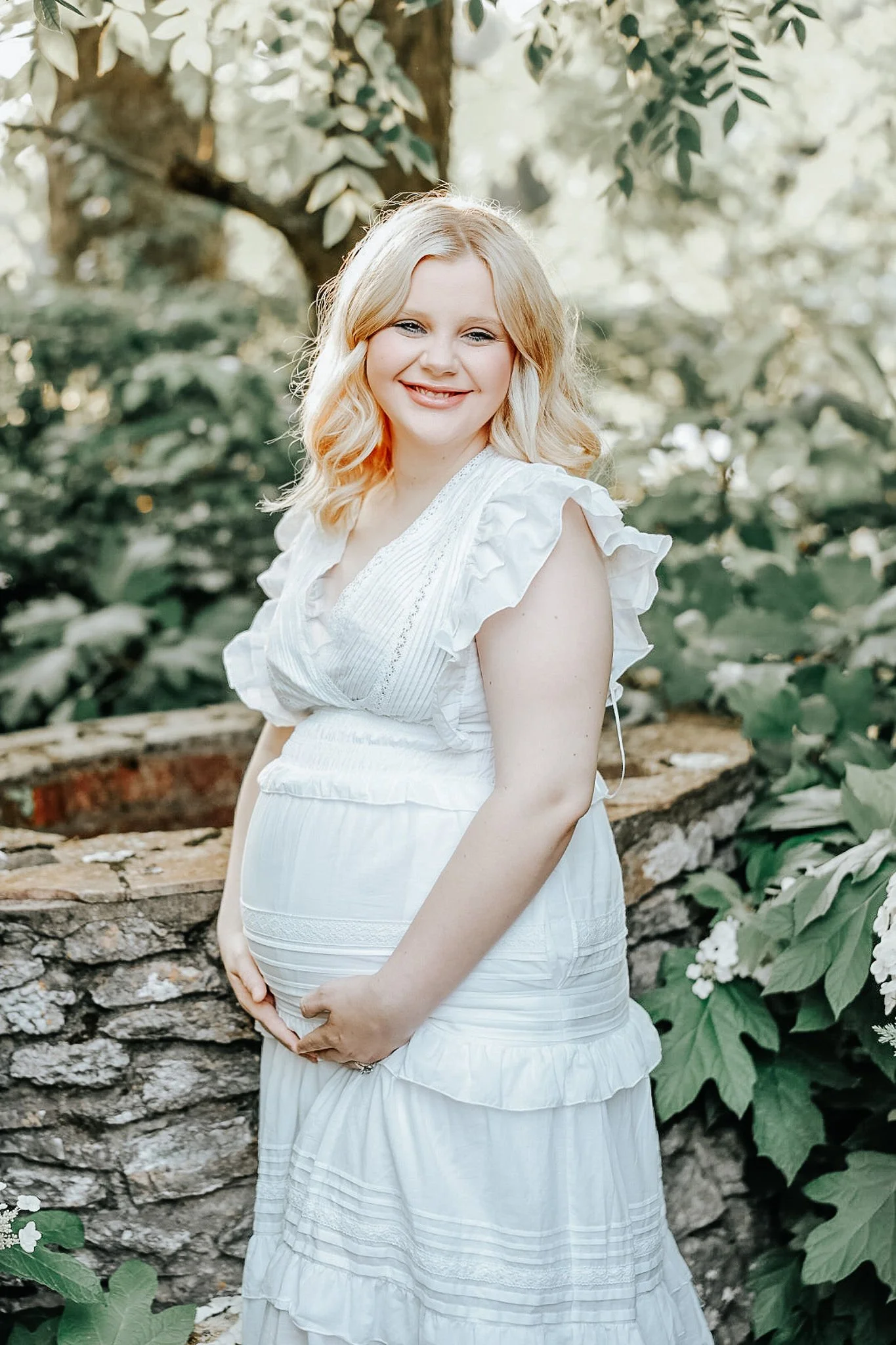 A pregnant woman with blonde hair in a white dress, standing outdoors amid greenery, smiling and gently holding her baby bump.