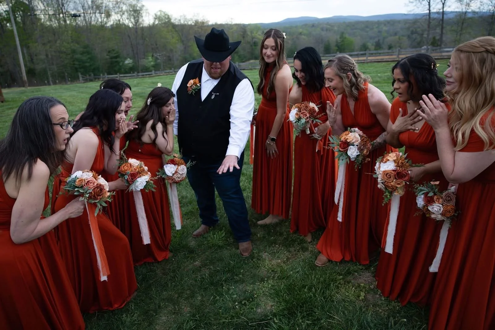 A group of women in matching red dresses holding bouquets, surrounding a man in a cowboy hat and vest, outdoors on grass with trees and mountains in the background, possibly at a wedding.