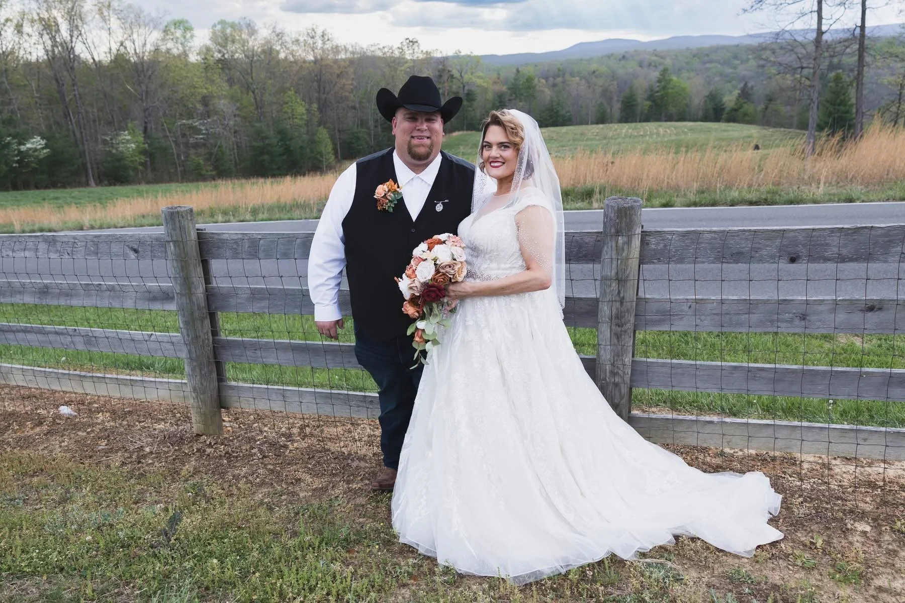 A bride and groom standing outdoors behind a wooden fence with a rural landscape of trees, grass, and mountains in the background. The bride is holding a bouquet of flowers, and both are smiling.