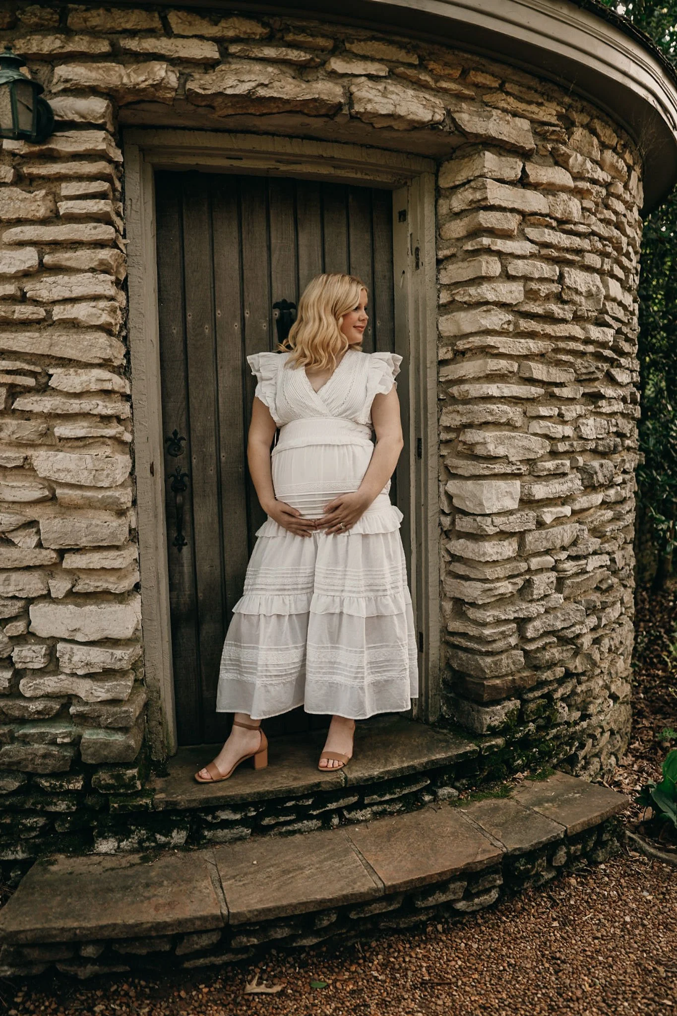 Pregnant woman in white dress standing on a stone stoop in front of a rustic wooden door, holding her belly and looking to her right.