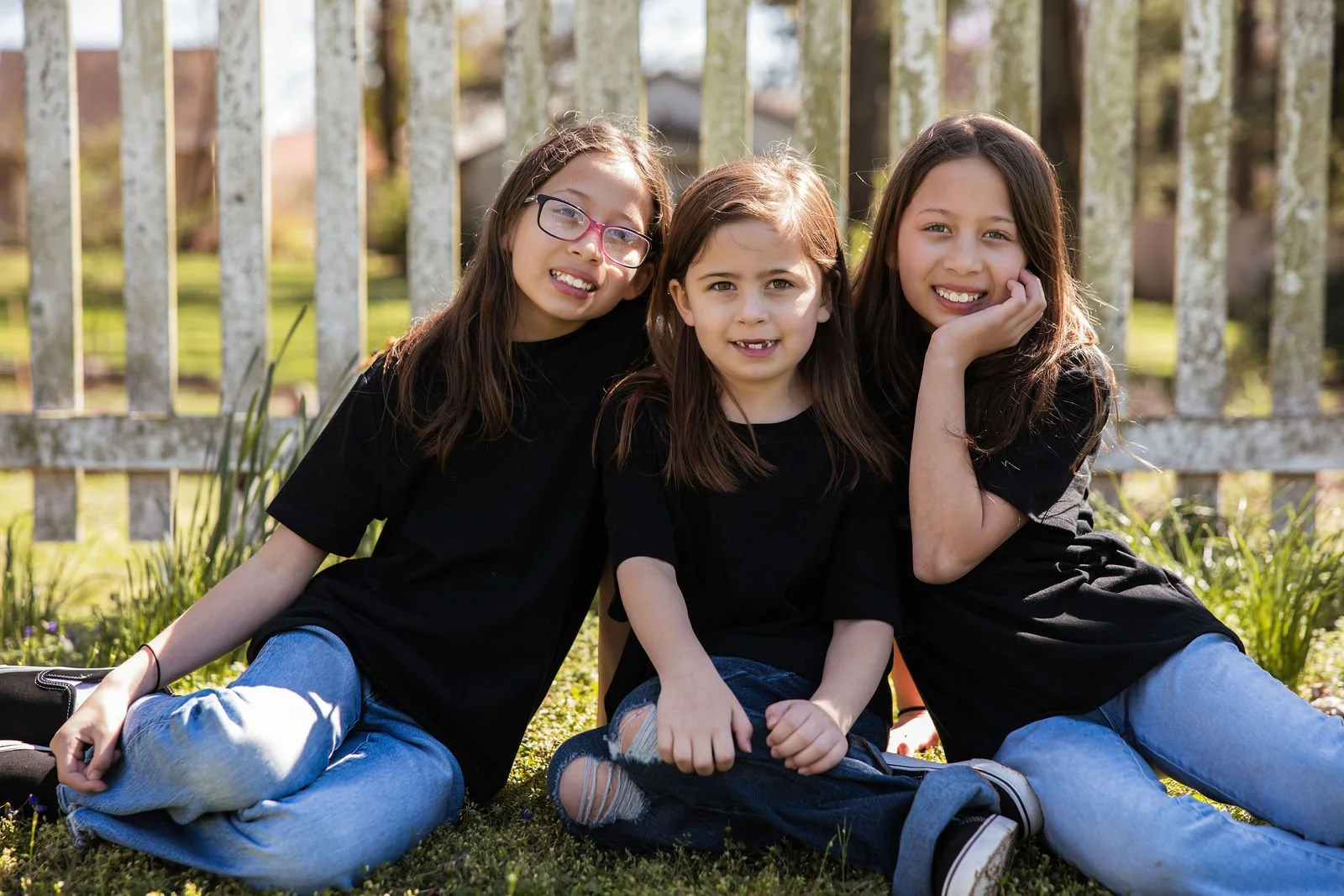 Three young girls sitting on grass in front of a white picket fence, smiling at the camera.