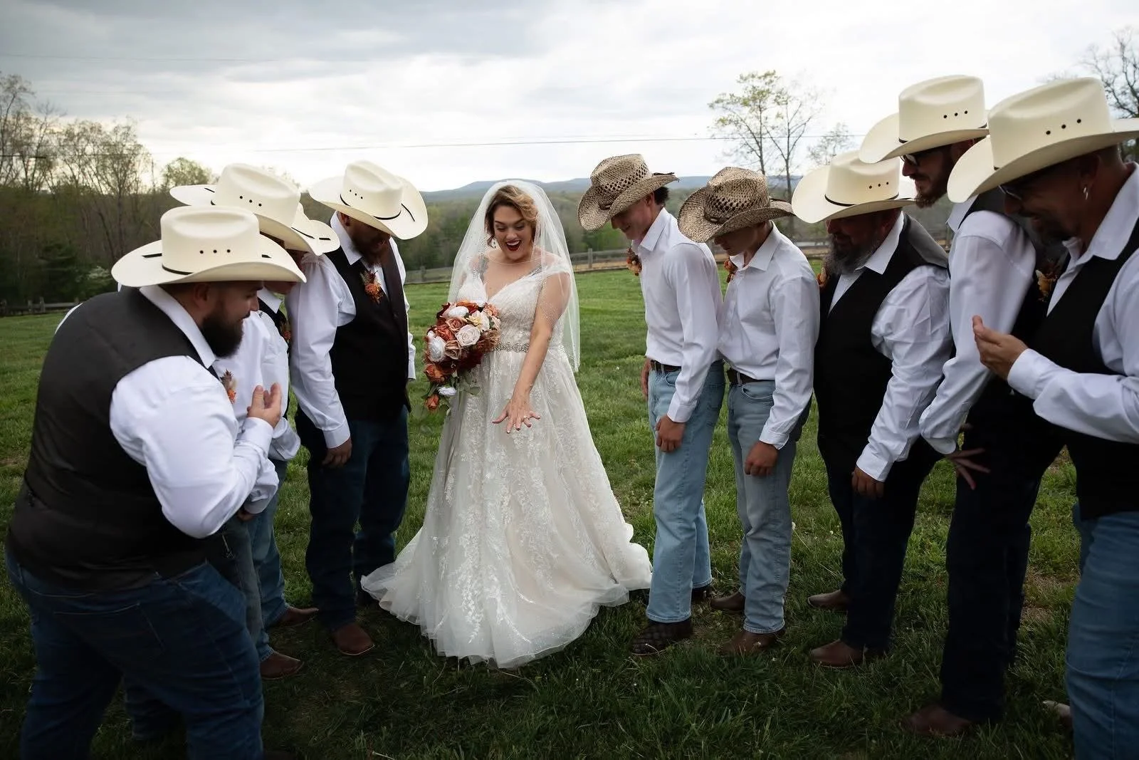 A bride and seven men in cowboy hats and western attire circle outdoors on grass, smiling and bowing their heads.