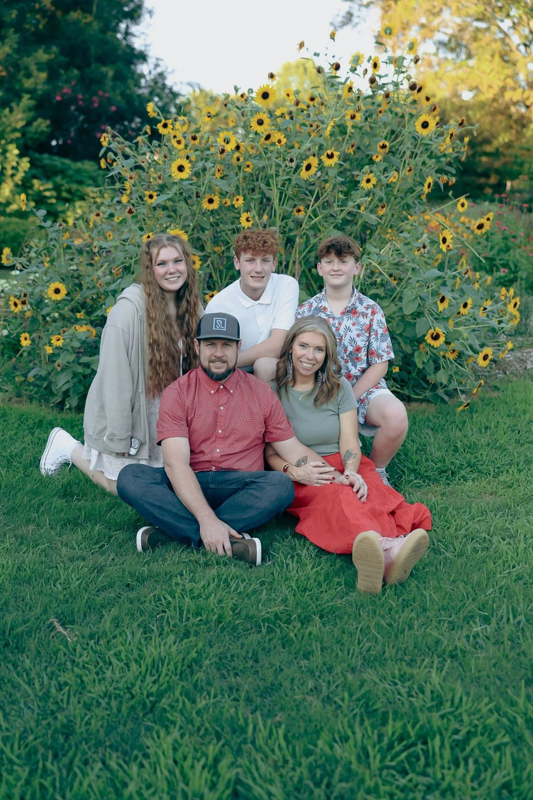 A family of five posing outdoors in front of a large sunflower plant, sitting on green grass during daytime.