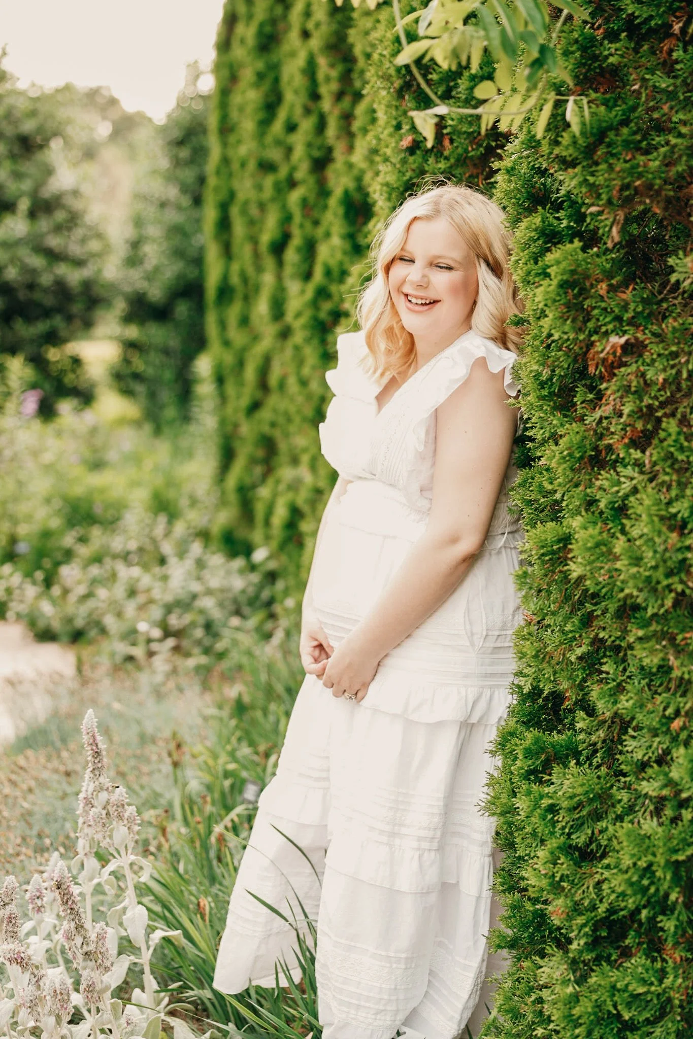 A woman with blonde hair smiling and leaning against a lush green hedge, dressed in a white sleeveless dress, in a garden setting.