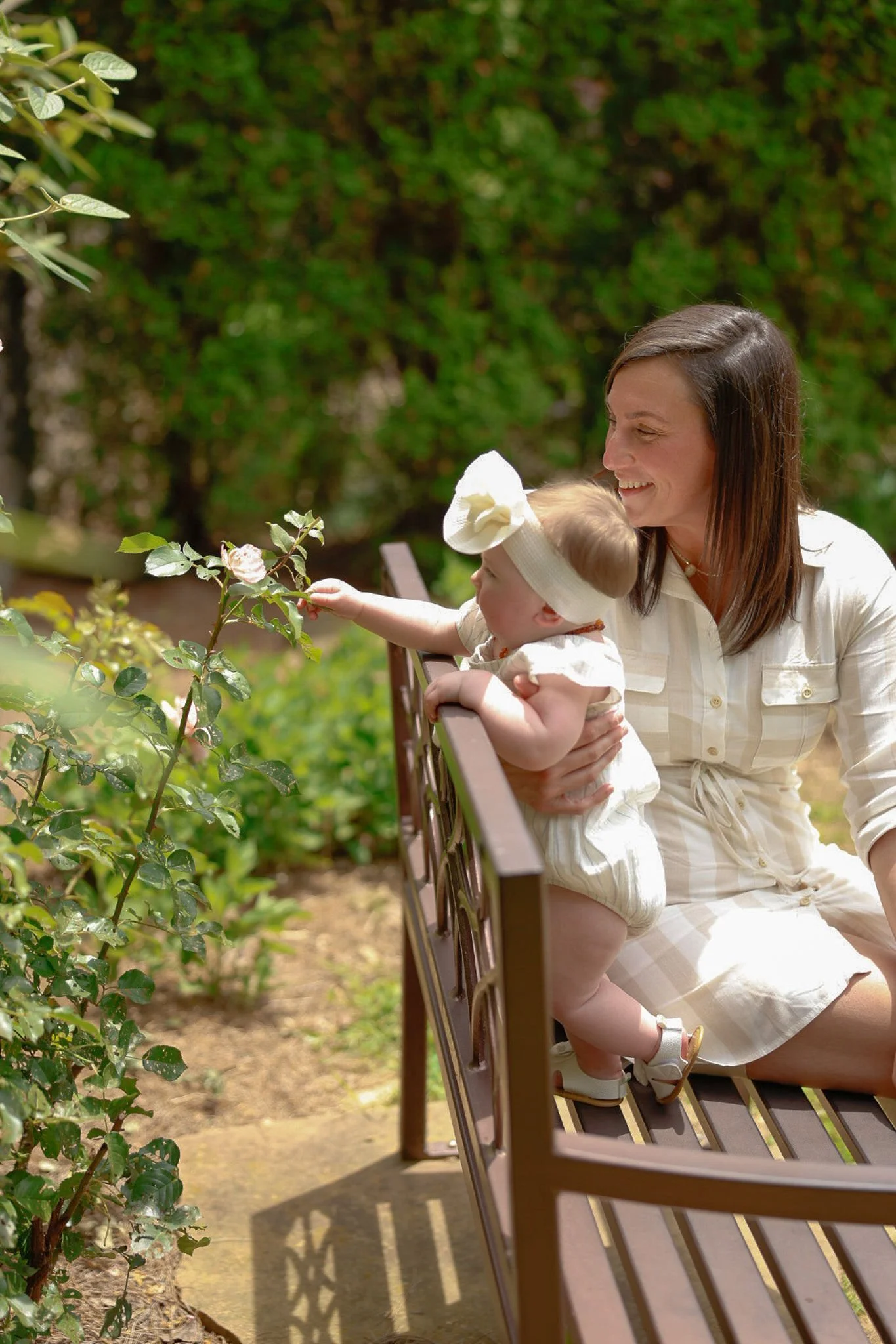 A woman and a young child sitting on a park bench outdoors, the woman holding the child as they look at and reach towards a flowering bush.