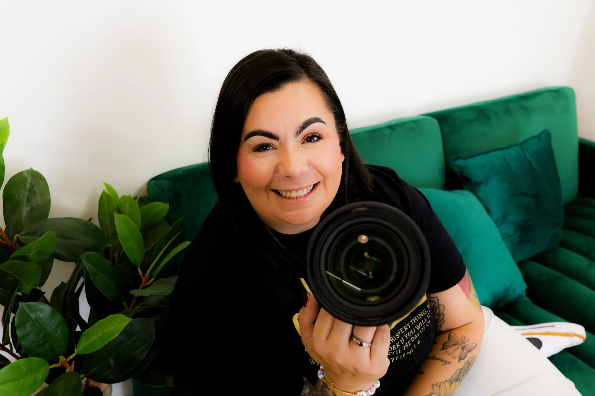 Smiling woman holding a professional camera lens pointing at the viewer, seated on a green couch with a green cushion, near a green plant.