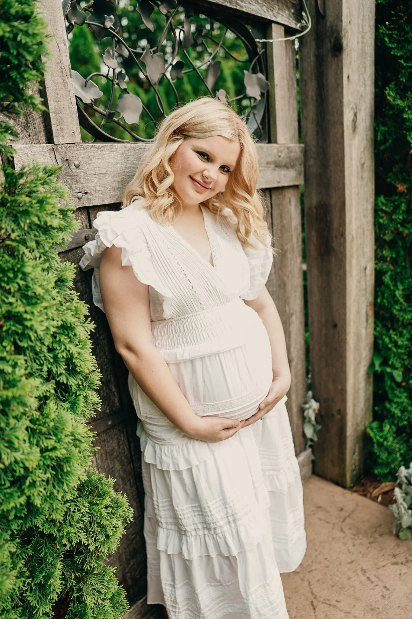 A pregnant woman with blonde hair in loose curls wearing a white ruffled dress, standing outdoors near a wooden gate and greenery, smiling and gently holding her belly.