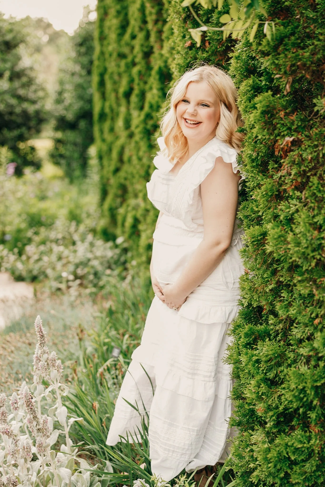 A woman with blonde hair, wearing a white dress, stands outdoors beside a lush green hedge, smiling and looking happy.