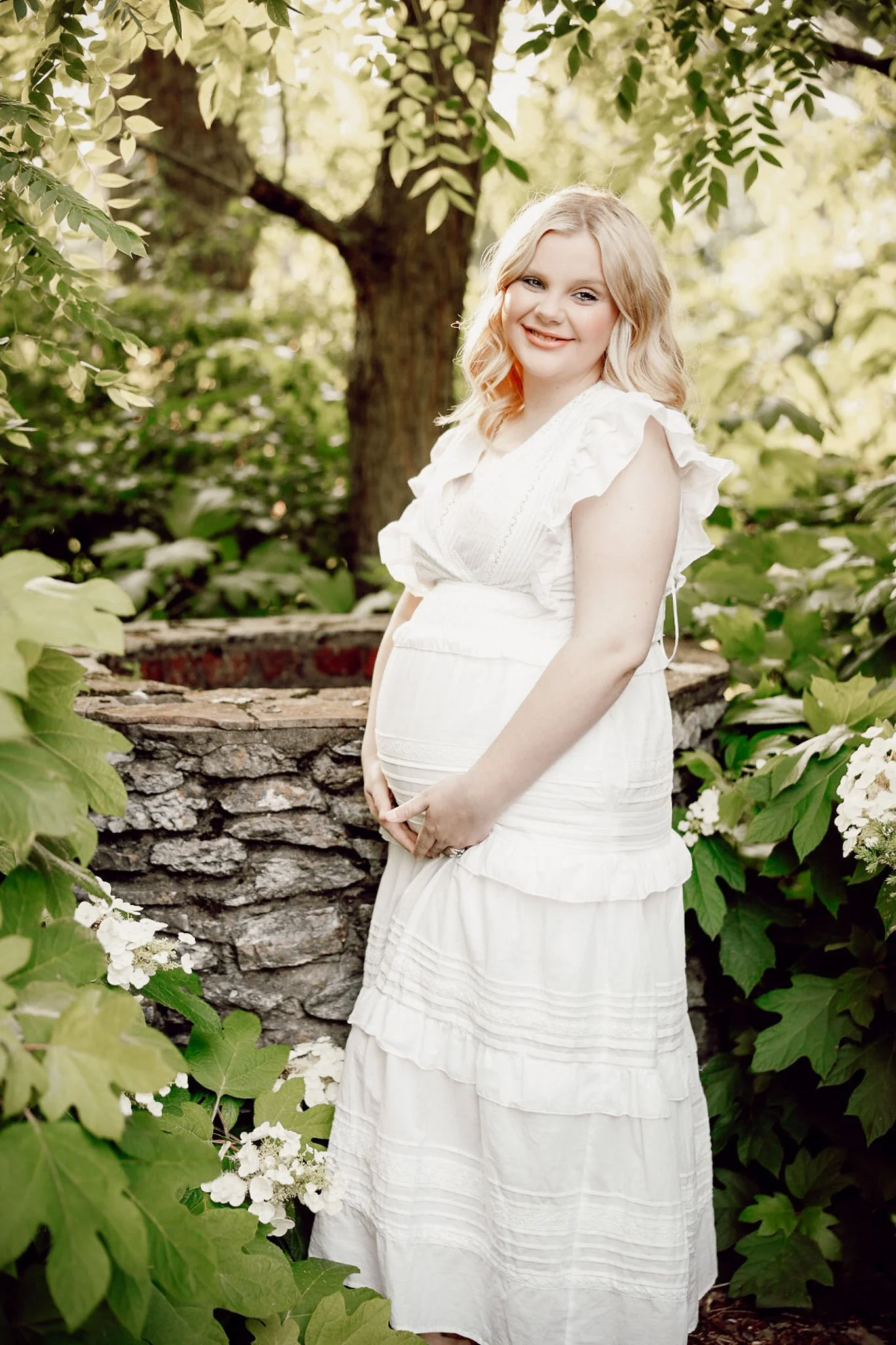 A pregnant woman with blonde hair, wearing a white dress, is smiling and standing outdoors amidst green foliage, with a stone well and tree in the background.