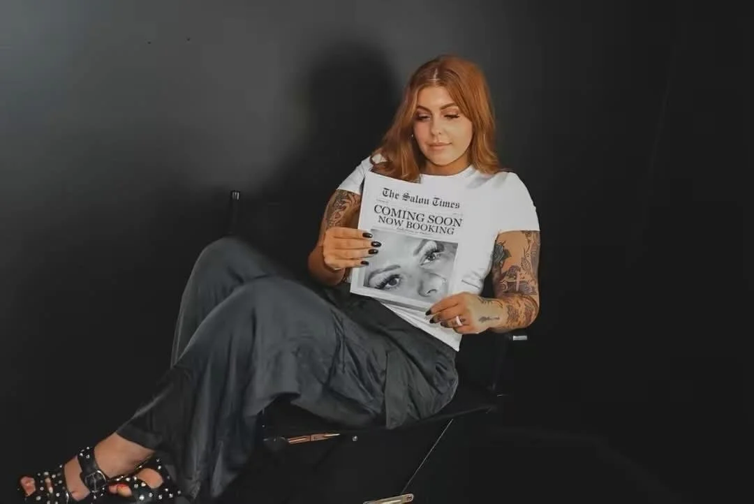 A woman with red hair and tattoos on her arms sitting on a black chair, looking at a newspaper that has a headline about The Salisbury Times with a black and white photo of a woman’s eyes.
