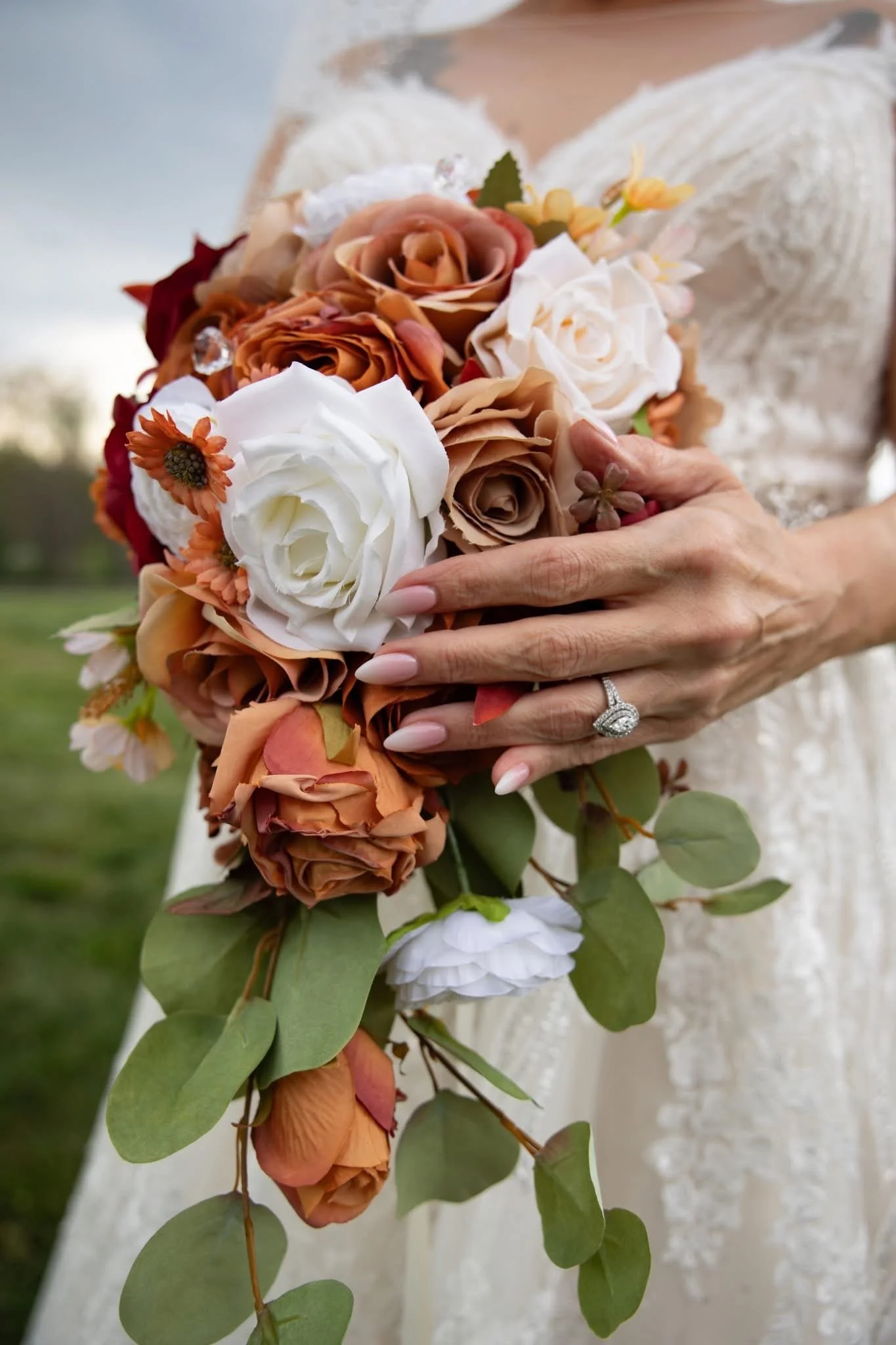 A person in a lace dress holding a bouquet of flowers with white, peach, and rust-colored roses, green leaves, and small orange flowers, outdoors in a park or garden setting.