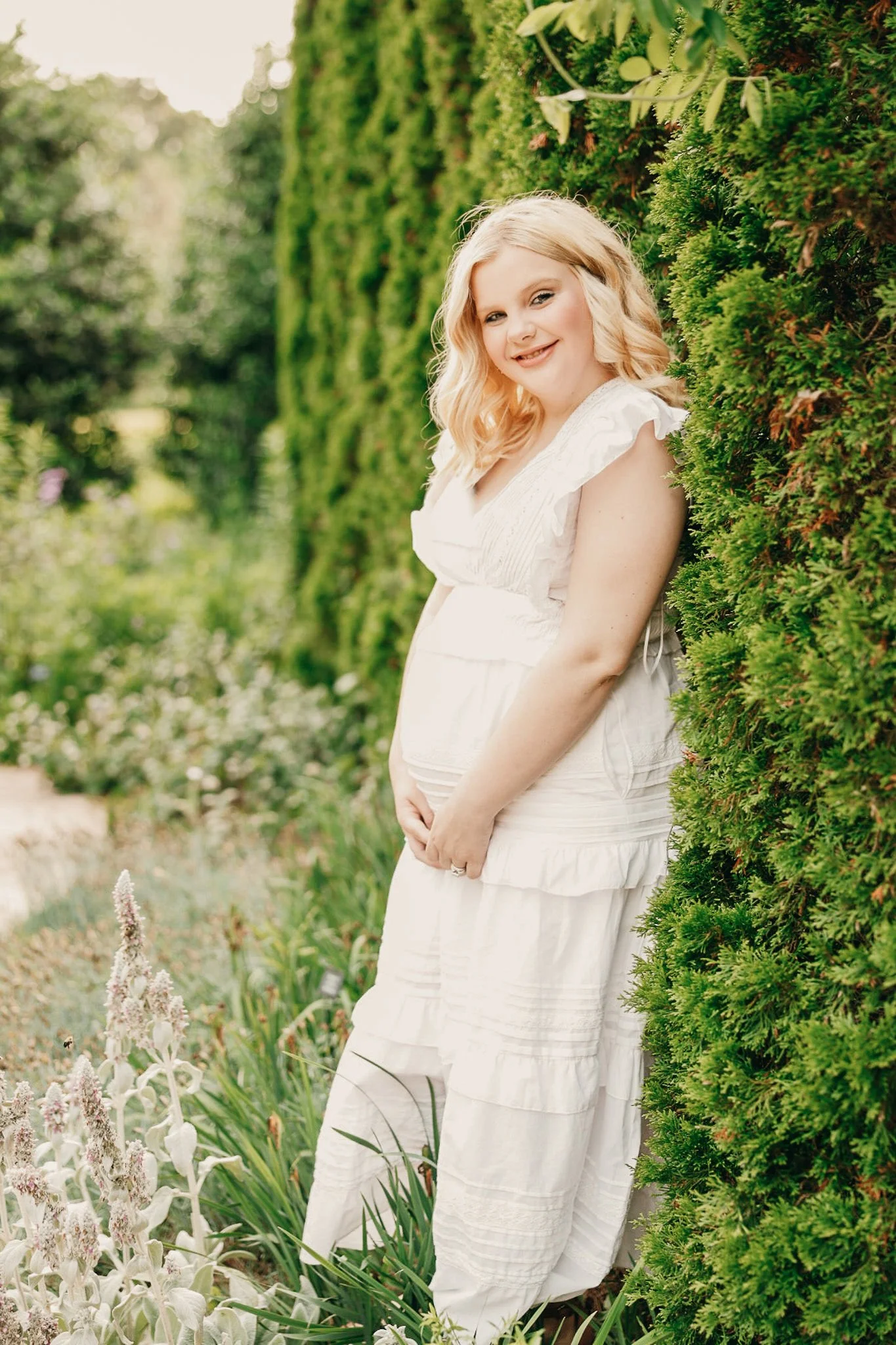 A young woman with blonde, wavy hair wearing a white dress, standing near a green hedge in a garden, smiling at the camera.