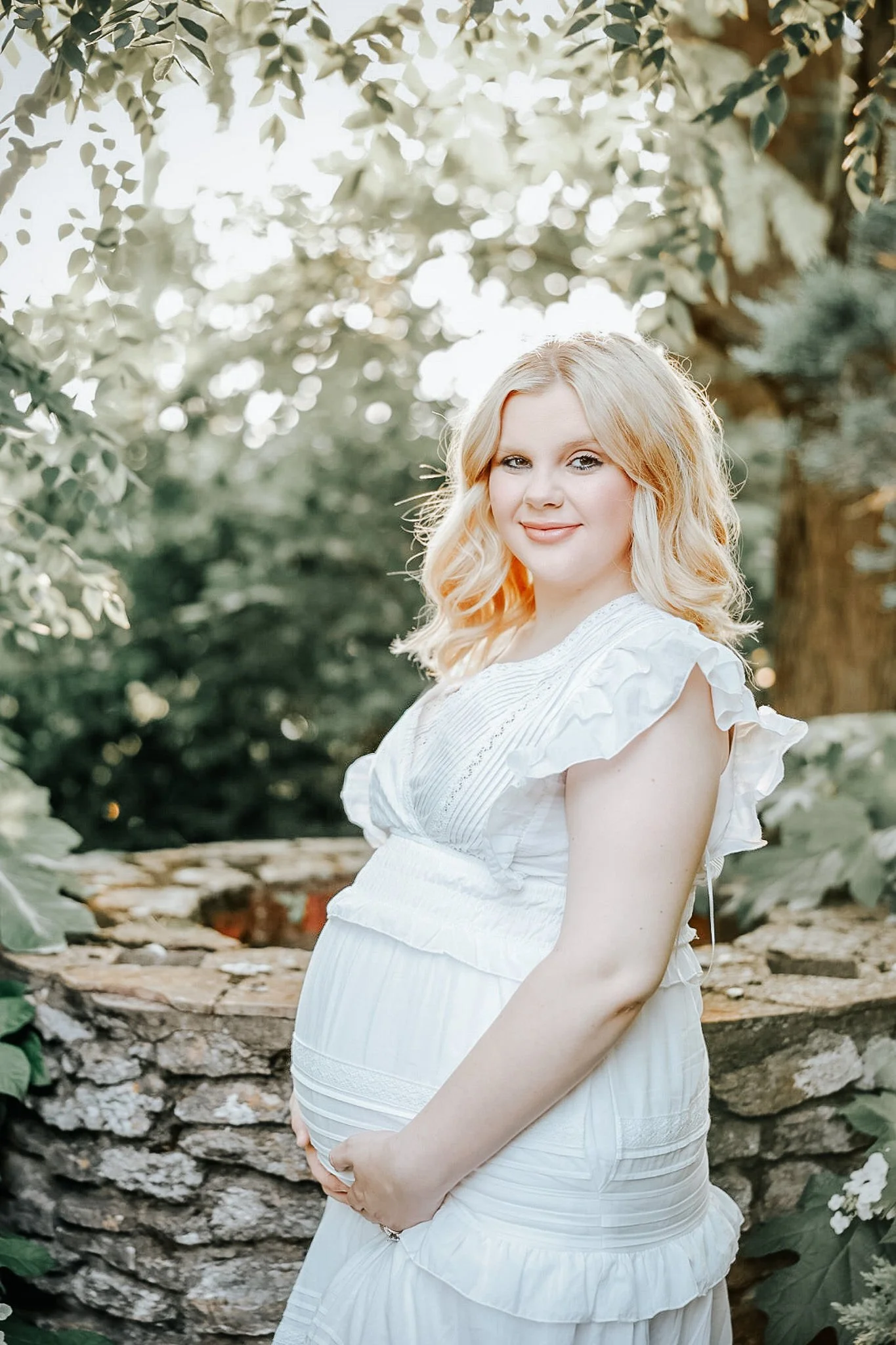 A pregnant woman with blonde hair standing outdoors near a stone well, wearing a white dress with ruffled sleeves, smiling softly at the camera.