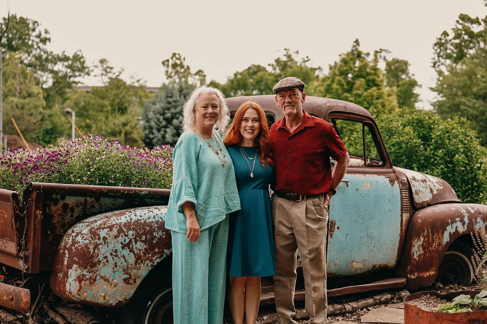 Three people, two women and one man, standing outdoors in front of an old, rusted truck with flowers in the bed, greenery, trees, and bushes in the background.