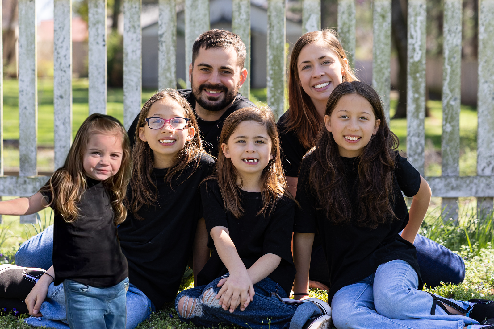 A family of six sitting and kneeling on grass in front of a white picket fence, smiling for the camera outdoors on a sunny day.