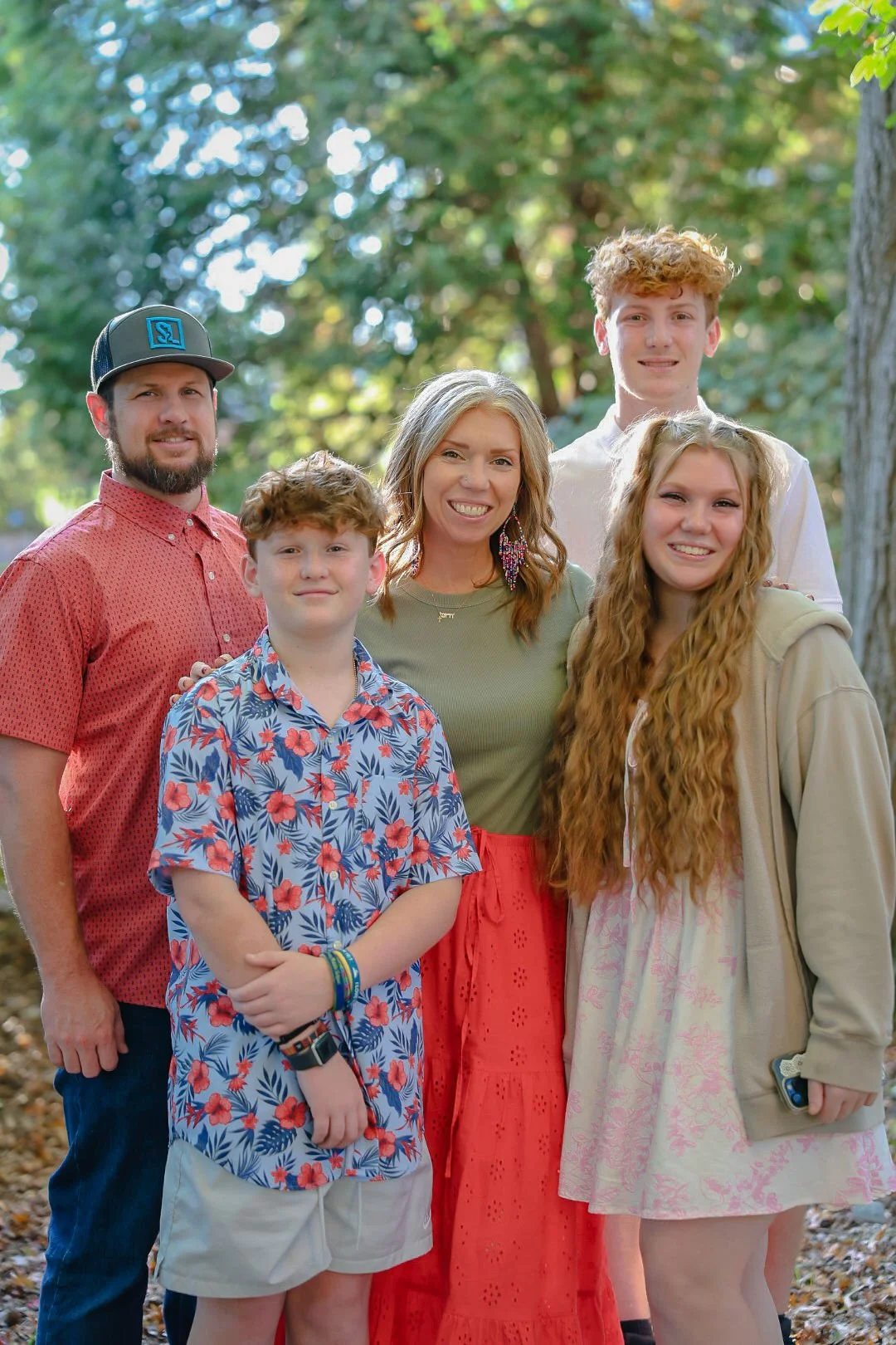 A smiling woman with long, curly hair standing outdoors with four children, in a wooded area with sunlight filtering through the trees.