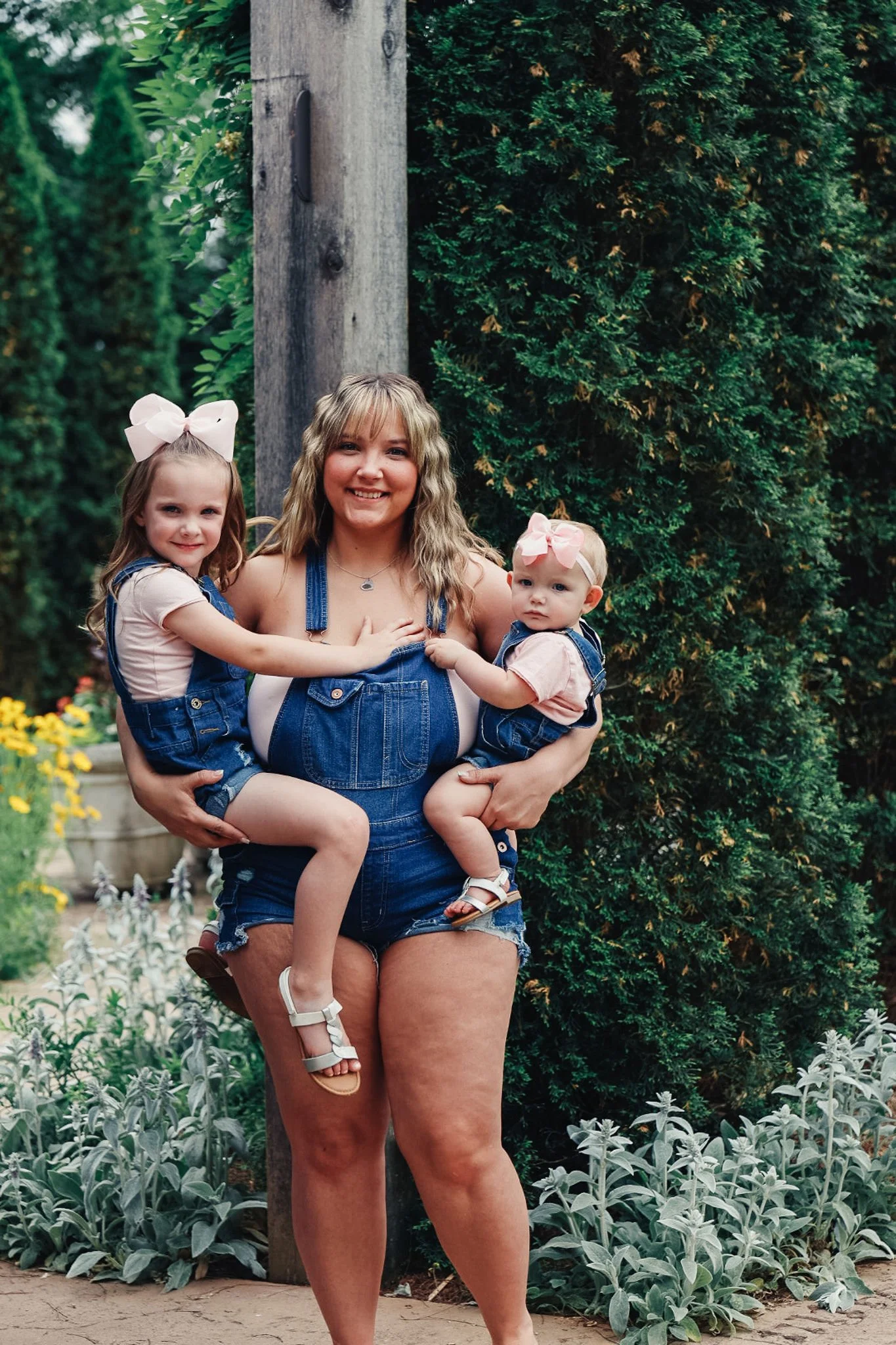 A smiling woman holding two young girls in a garden. The woman and girls are wearing denim overalls and pink bows, surrounded by greenery and flowers.