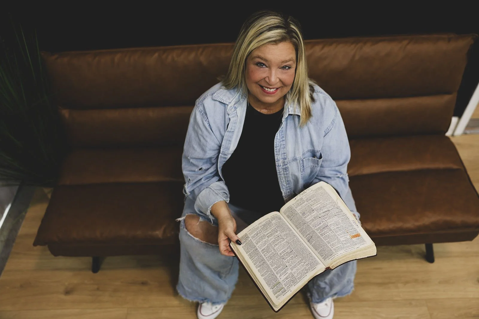 A smiling woman with blonde hair, wearing a black shirt and ripped denim jacket and jeans, sitting on a brown leather couch, holding an open large Bible, looking up at the camera.