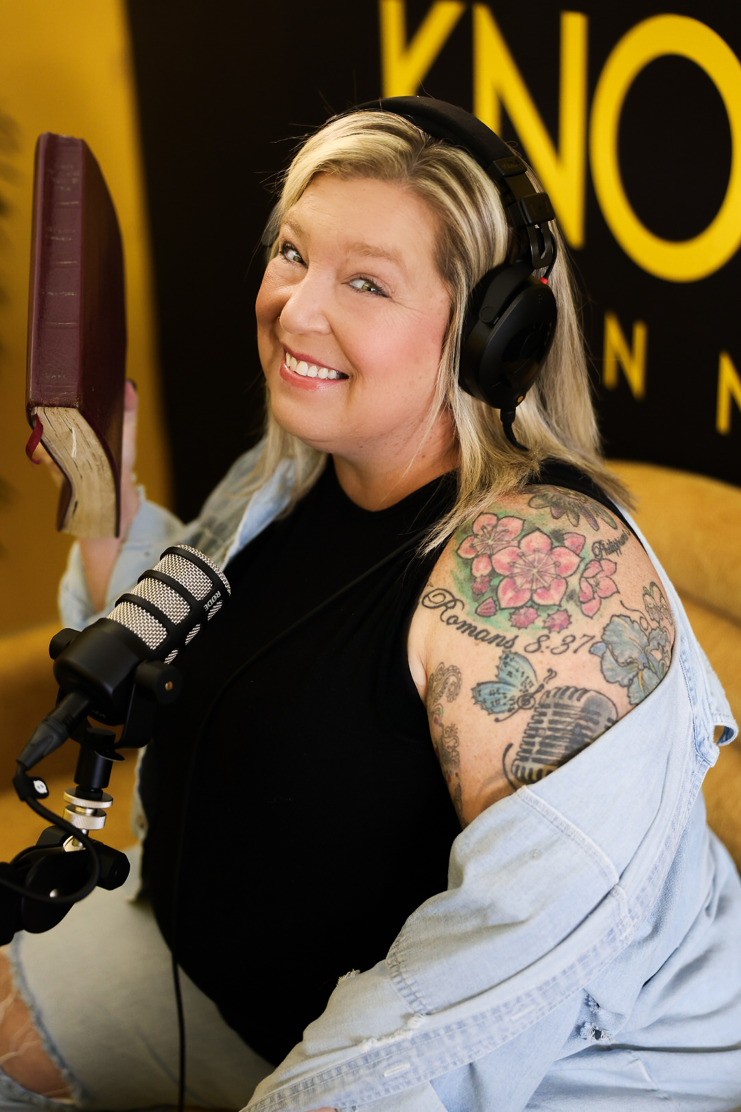 A woman with blonde hair, tattoos on her arm, and a big smile, wearing headphones and holding a book, sitting in front of a microphone in a radio studio.