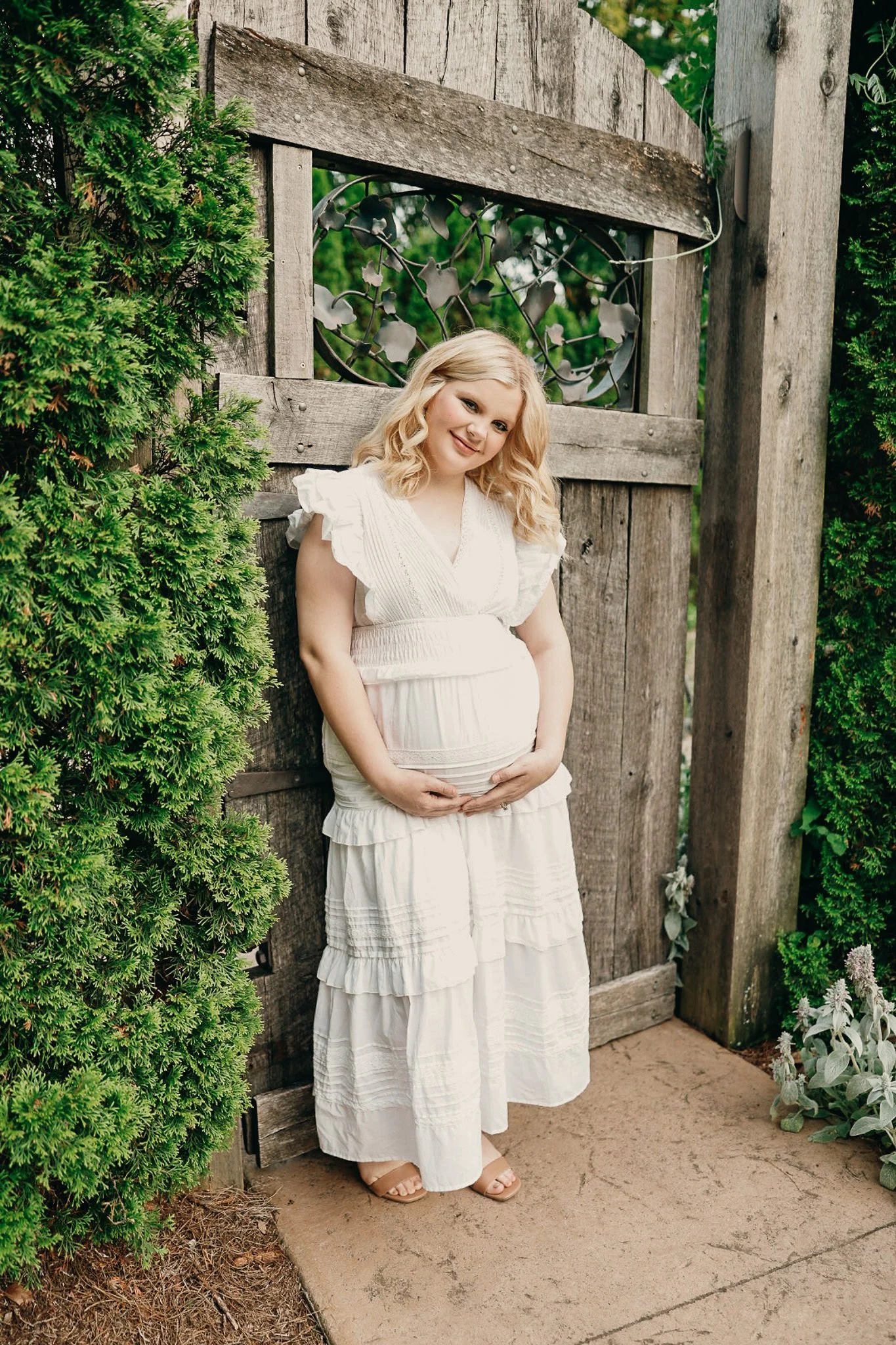 Pregnant woman standing outdoors against a rustic wooden gate, holding her belly, dressed in a white flowing dress, surrounded by greenery.