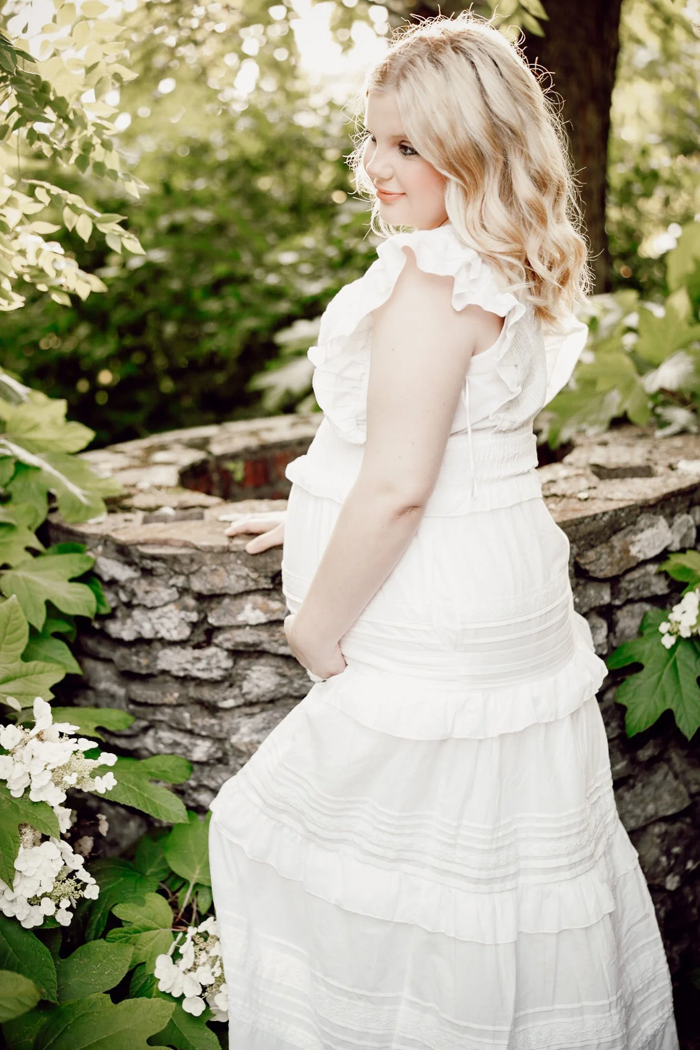 A young woman with blonde curly hair standing outdoors near a stone well and surrounded by green leafy plants and white flowers, wearing a white dress with ruffled sleeves.