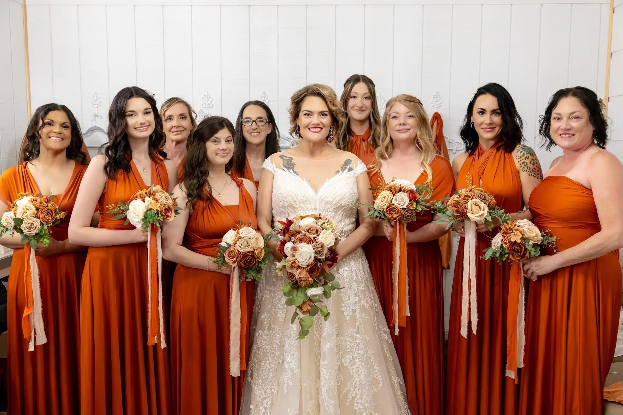 A bride and her bridesmaids standing together, all holding bouquets, in a room with white walls. The bride is in a white lace wedding dress, and the bridesmaids are in matching burnt orange dresses.