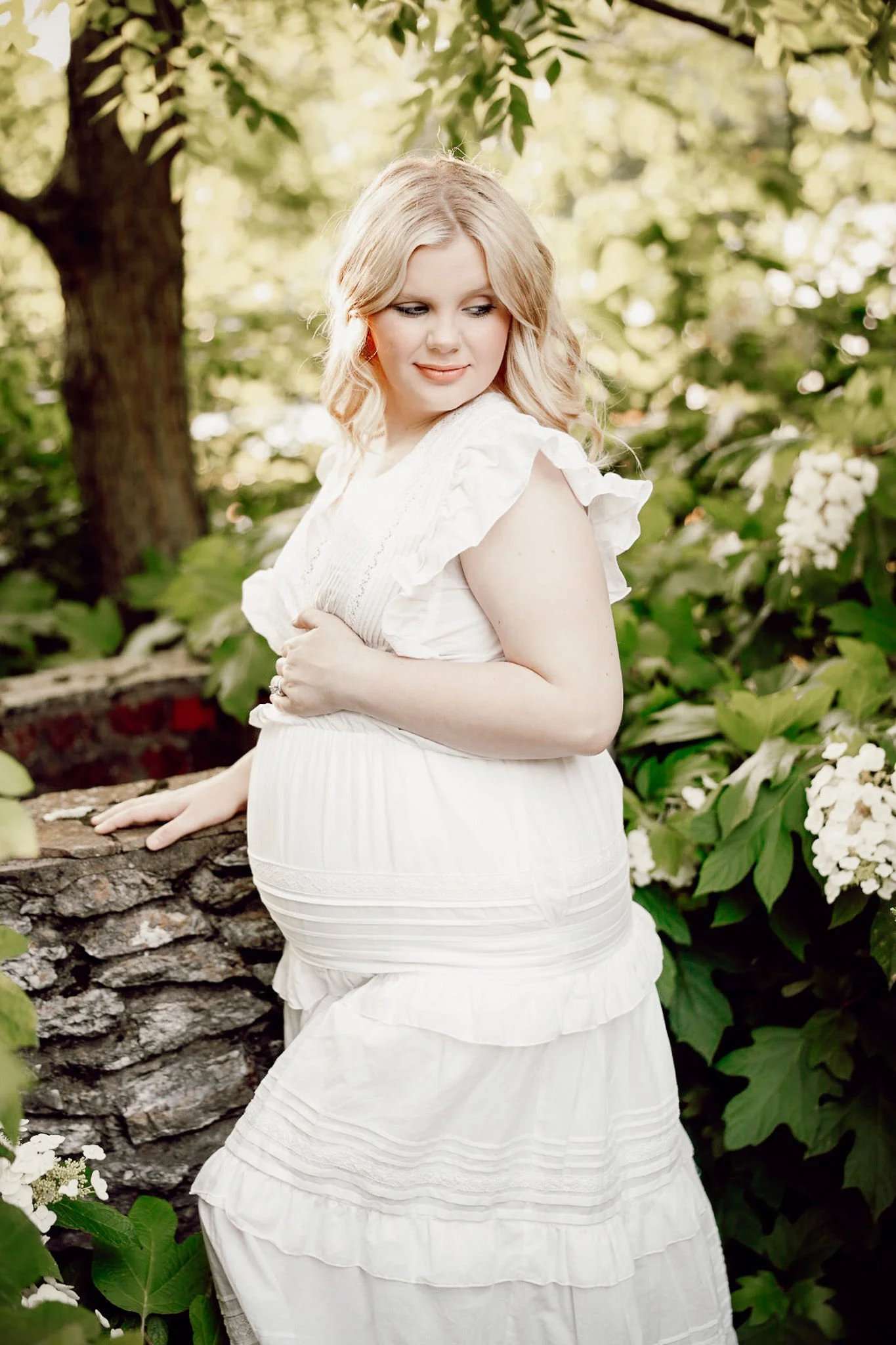 A pregnant woman in a white dress standing outdoors near bushes and trees with white flowers, touching a stone wall.