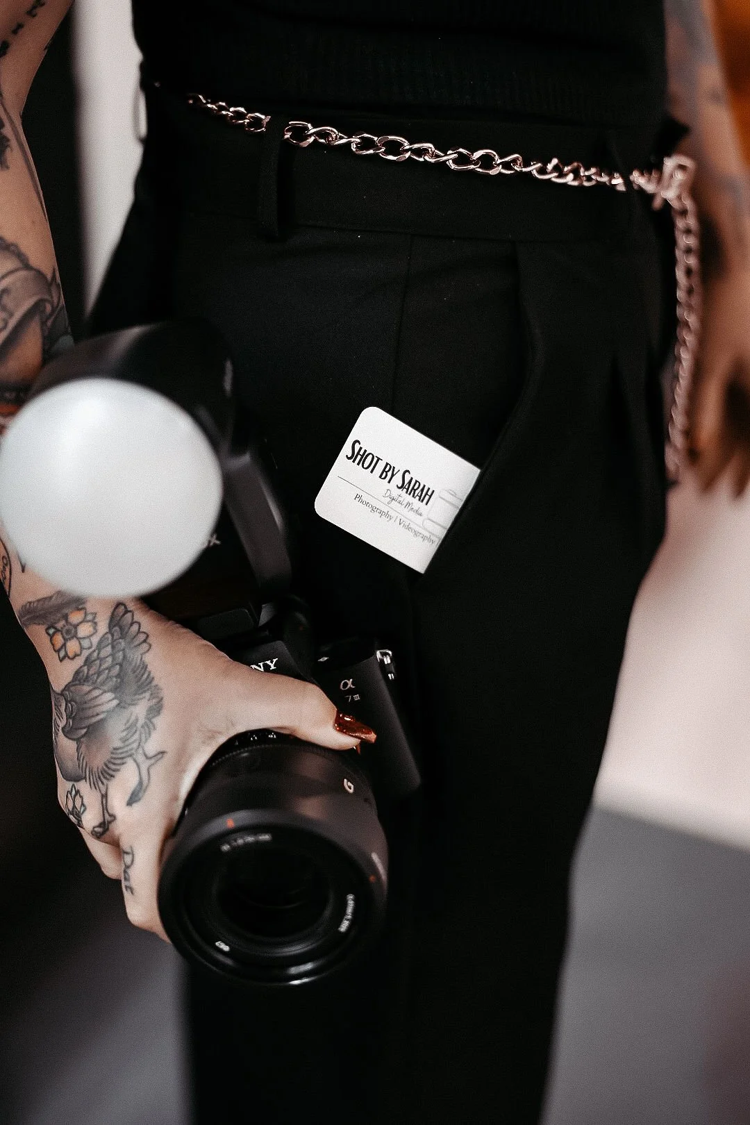 Close-up of a photographer wearing black pants and a chain belt, holding a camera, with a tattooed hand. A name tag reads 'Shot by Sarah'.