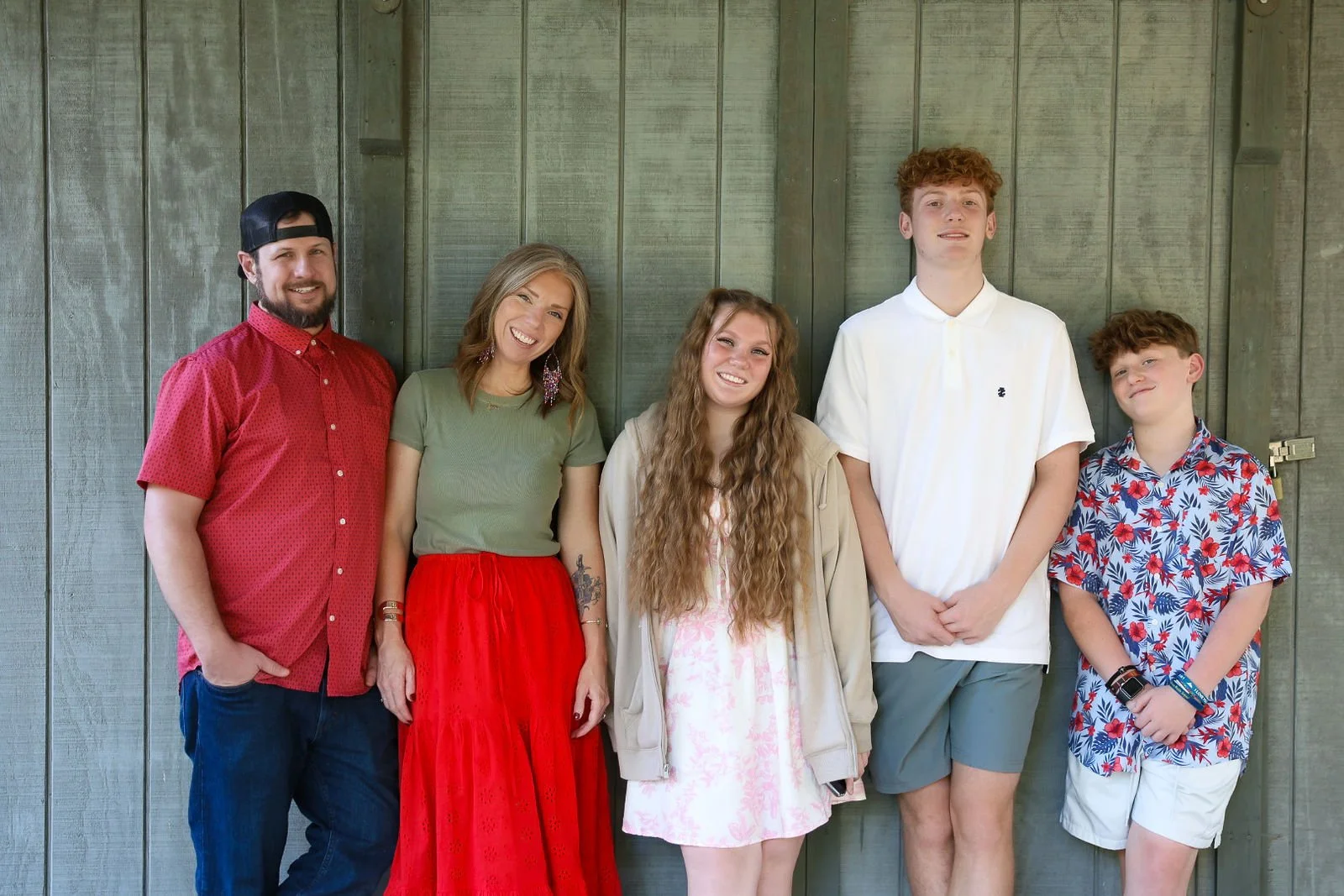 Group of six people standing against a wooden wall, smiling at the camera. Two women and four men, dressed casually in colorful clothing.