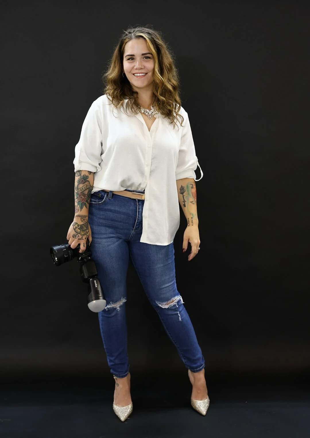 A woman with tattoos, wearing a white shirt, jeans, and high heels, holding a camera and standing against a black background.
