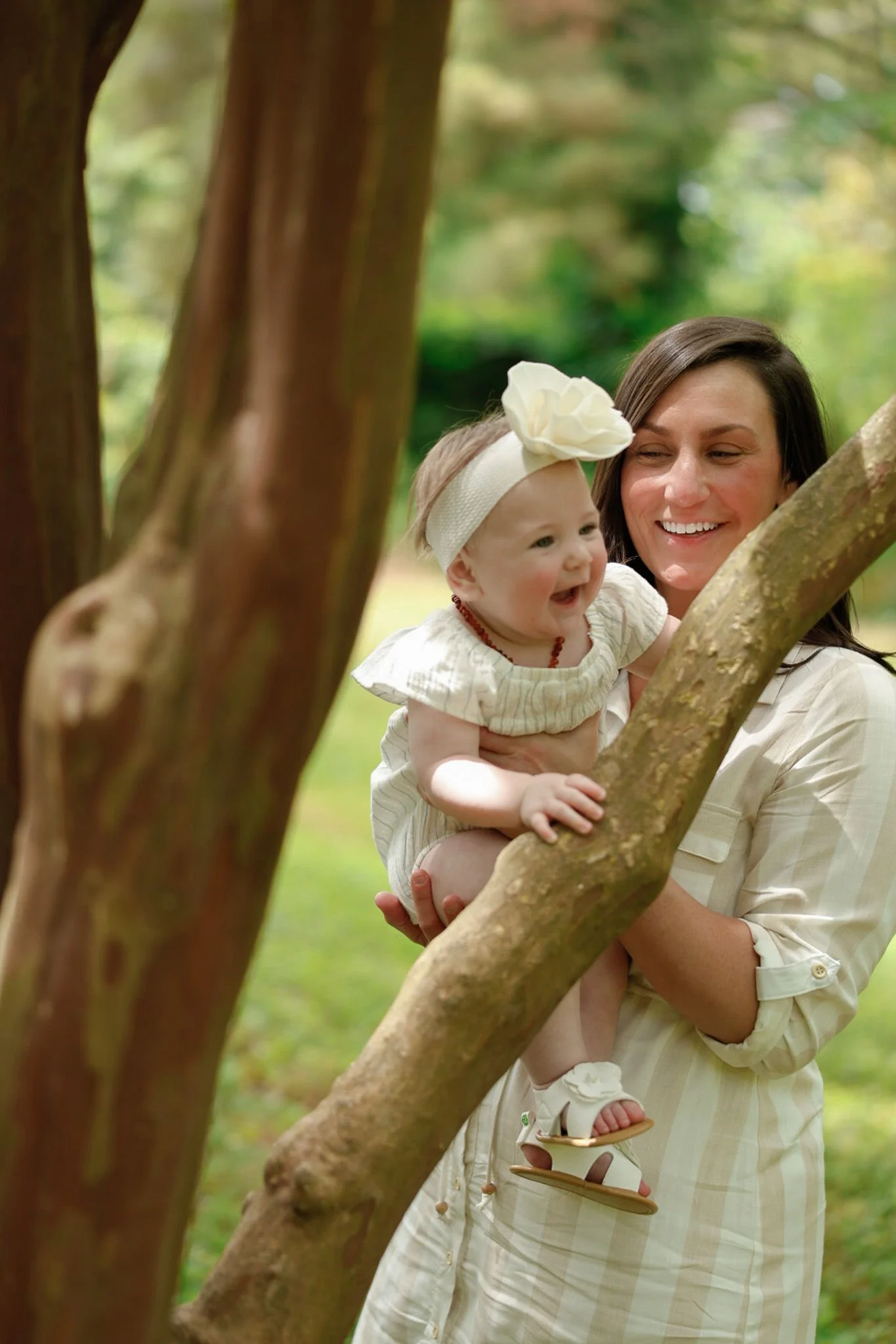 A woman holding a smiling baby girl on a tree branch outdoors, surrounded by greenery.