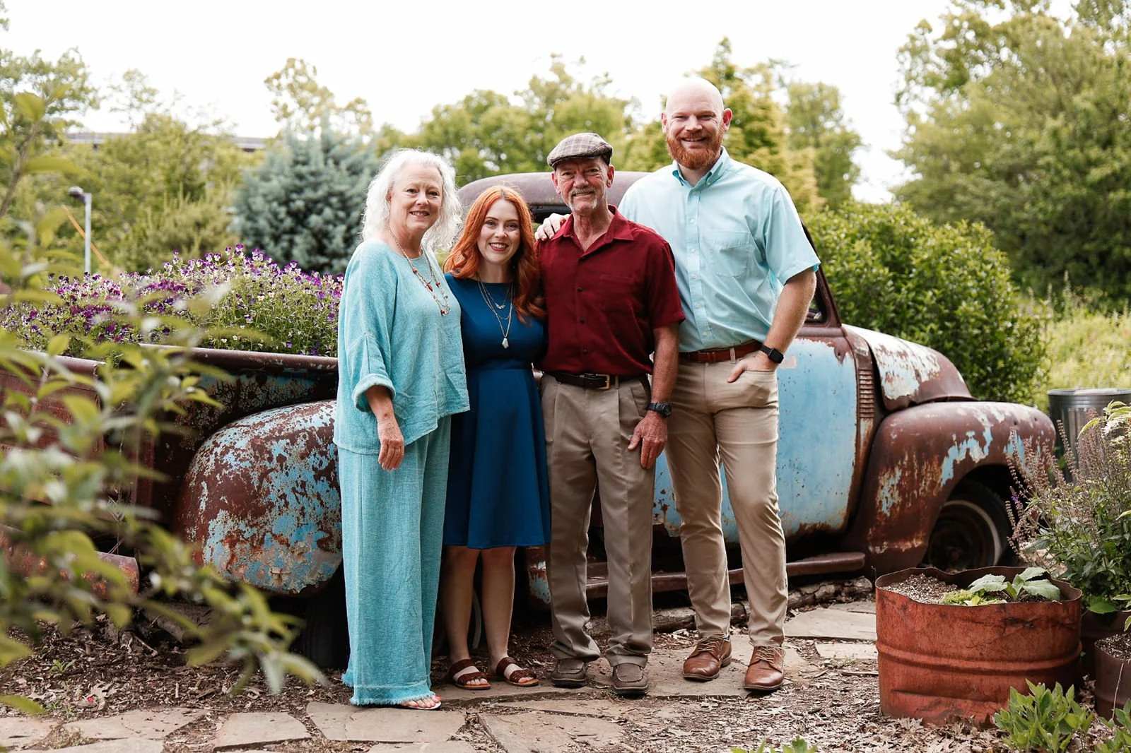 Family of five posing outdoors in a garden with a rusty, vintage truck behind them. The group includes two elderly women, two middle-aged men, and a young woman with red hair, all smiling.