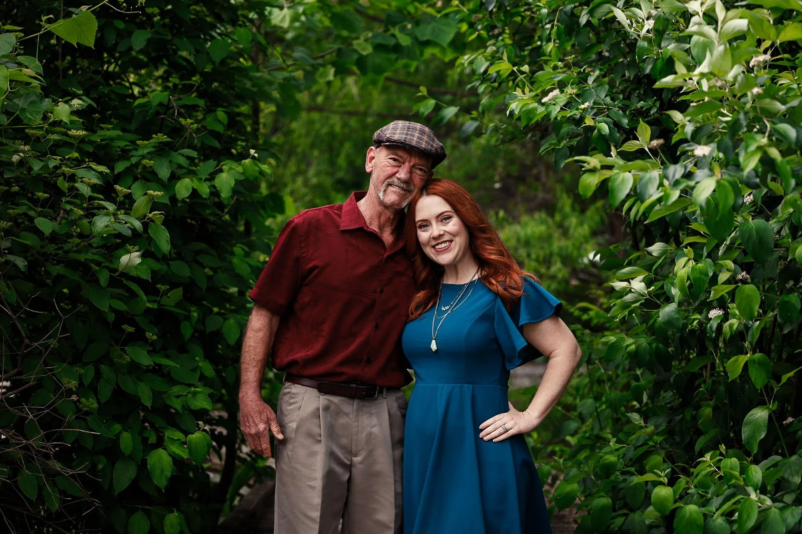 A man and woman standing close together outside amidst green shrubbery, smiling at the camera.