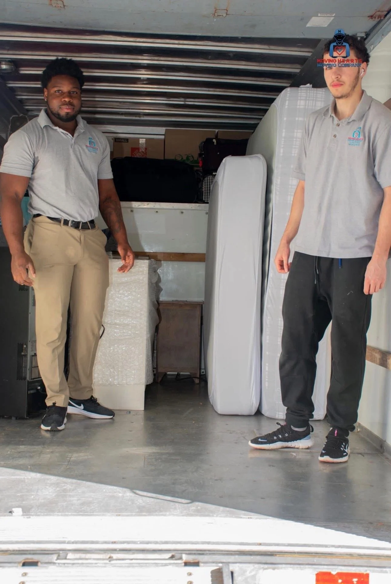 Two men standing in a truck loaded with furniture and boxes, wearing matching gray polo shirts with a moving company's logo.