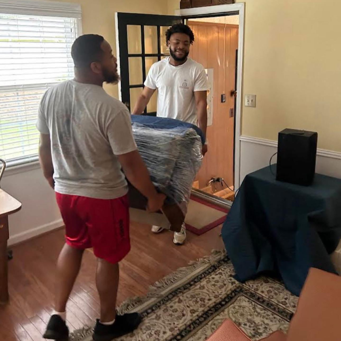 Two men are moving a large wrapped object into a house through a doorway, with a woman watching inside. The man outside is wearing a gray t-shirt and red shorts, and the man inside is wearing a white t-shirt.
