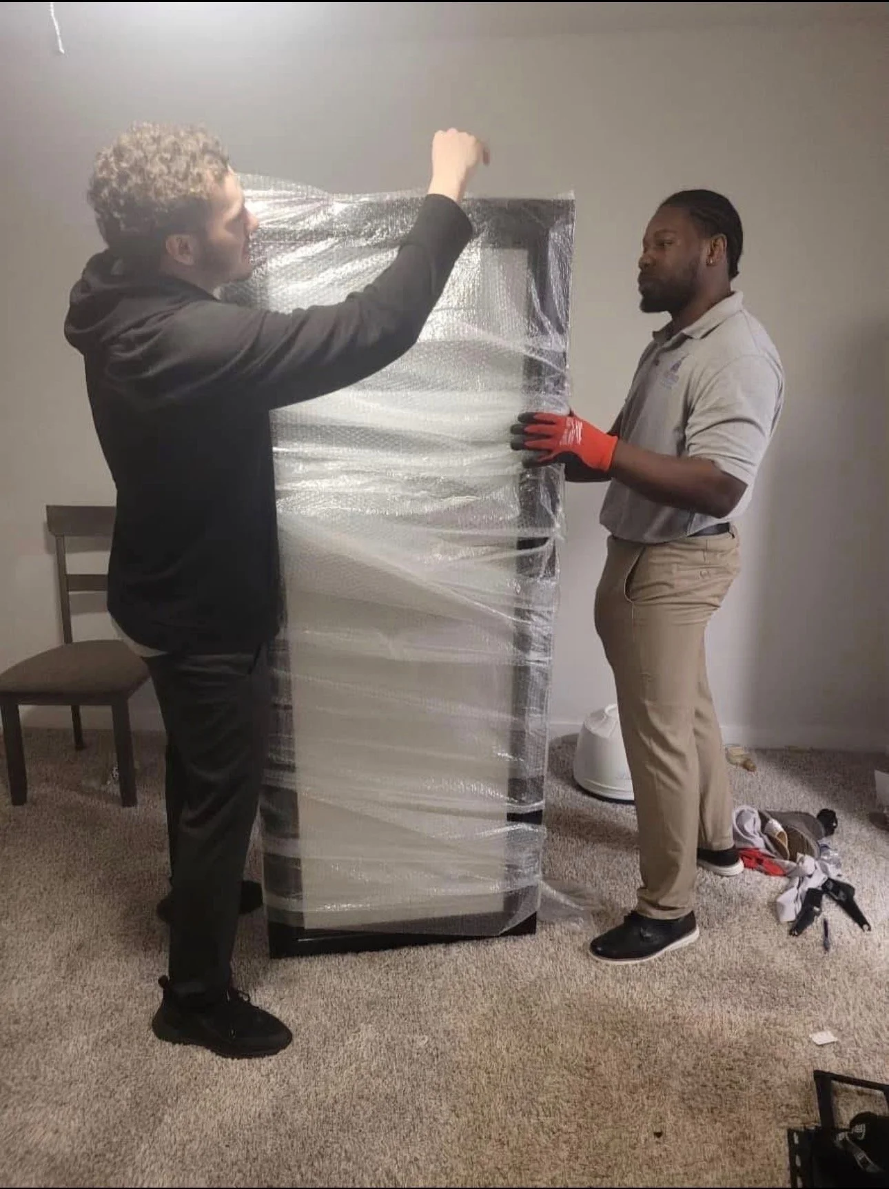 Two men unwrapping and preparing to move a wrapped refrigerator, using plastic wrap to secure it, in a room with beige carpet and plain white walls.