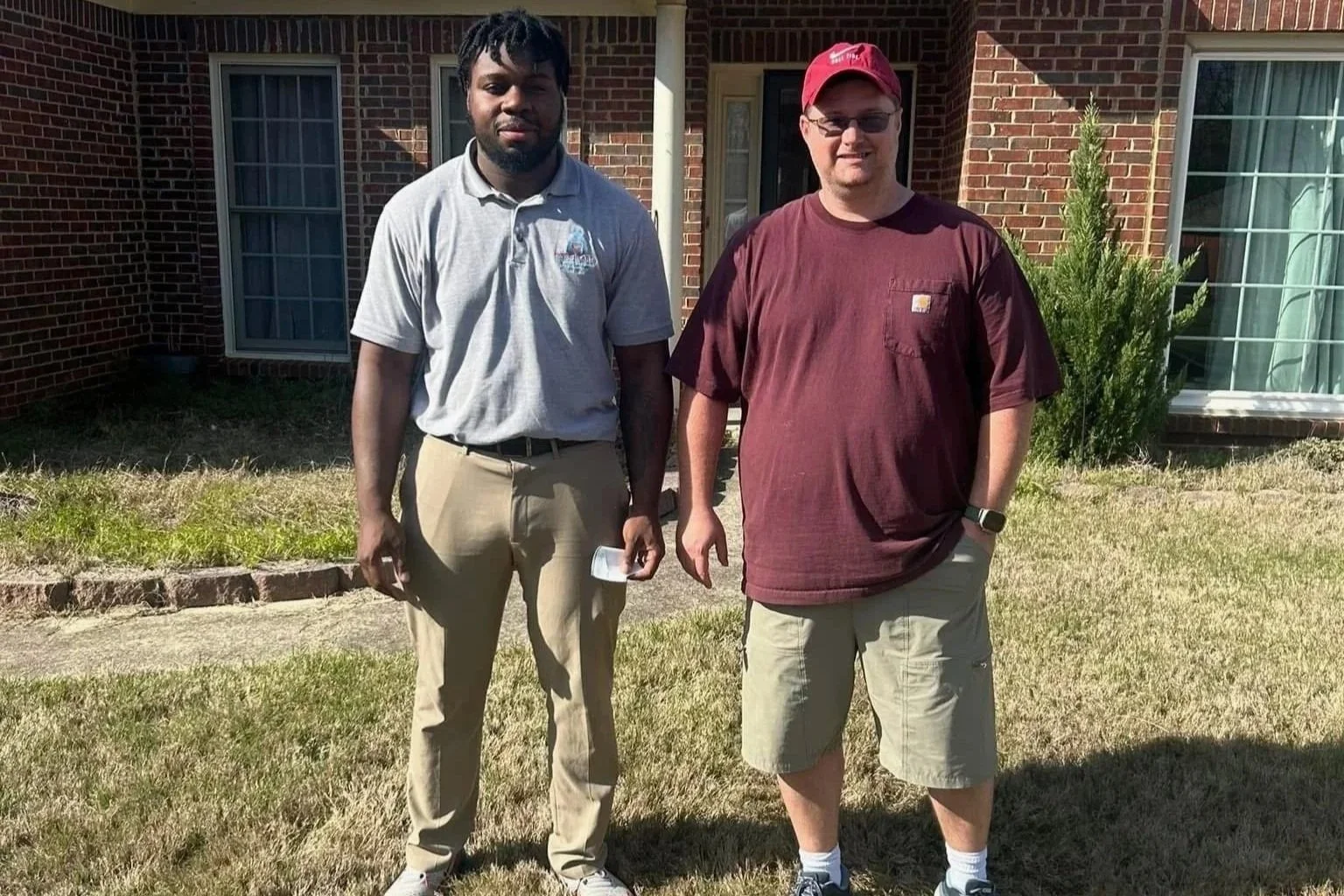 Two men standing outside in front of a brick house, one wearing khakis and a gray polo shirt, the other in khaki shorts and a maroon T-shirt, with a small shrub and window behind them.