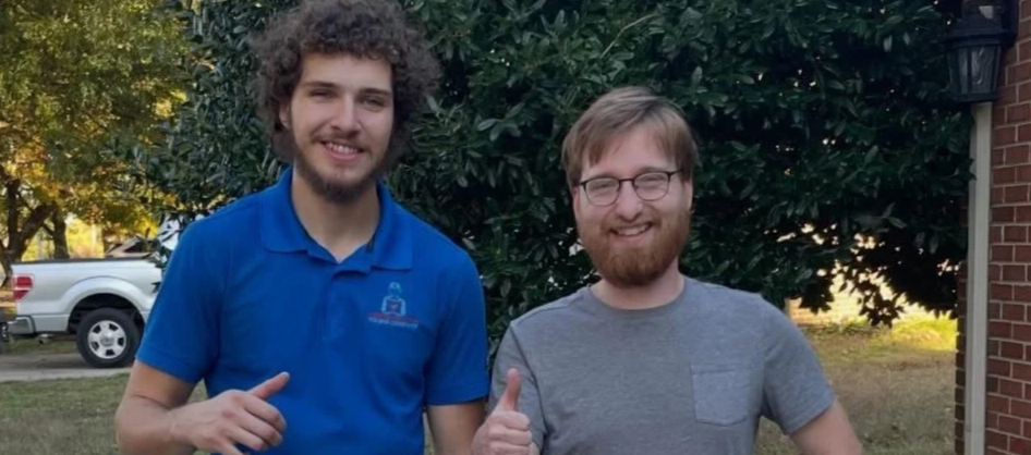 Two young men standing outdoors smiling, one in a blue polo shirt and the other in a gray t-shirt, with trees and a brick building in the background.