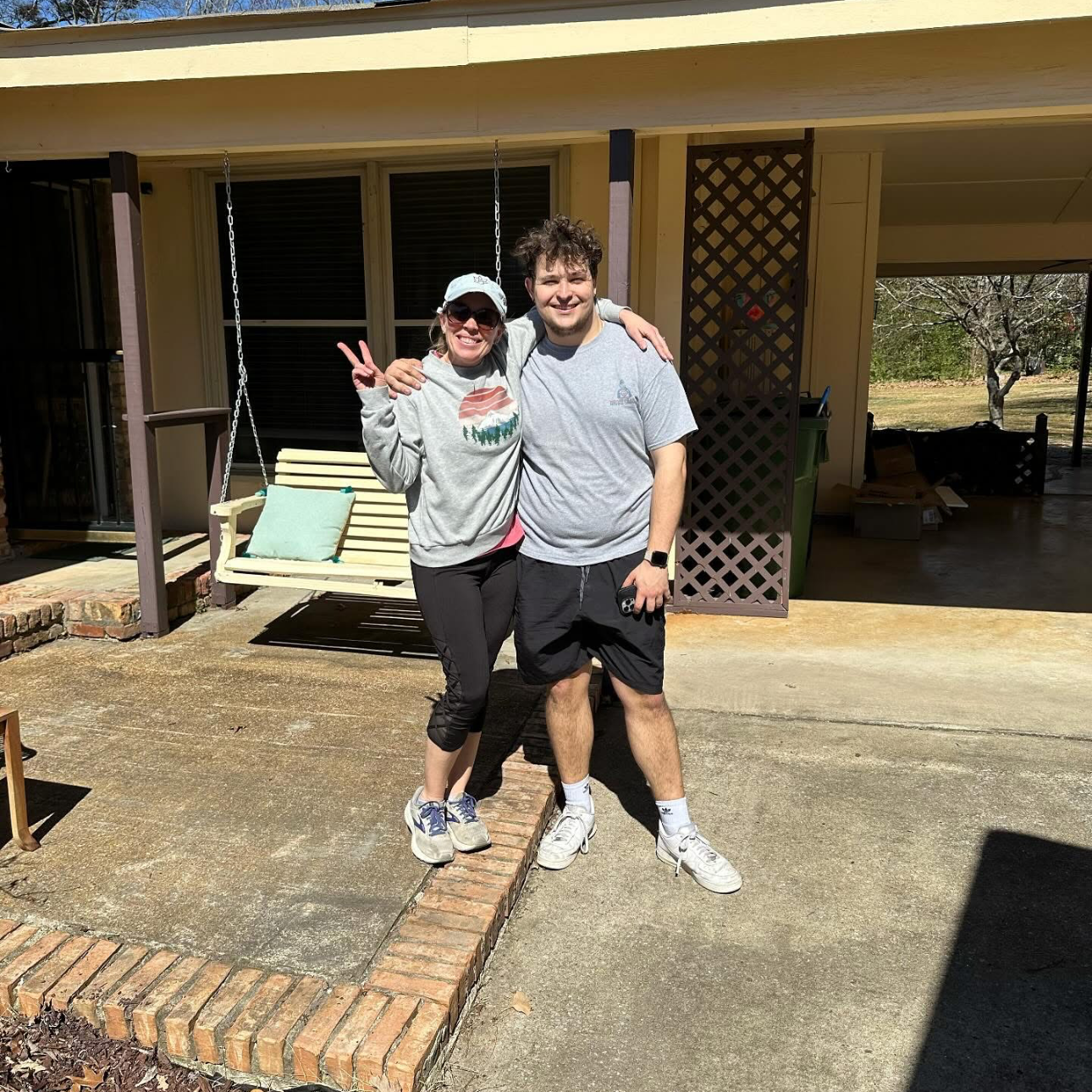 Two people standing on a porch, smiling for the camera. The woman on the left is making a peace sign, wearing sunglasses and a gray sweatshirt. The man on the right is wearing a gray T-shirt and shorts, holding a small object in his left hand. Behind them are porch swings and a house with open doors and windows.