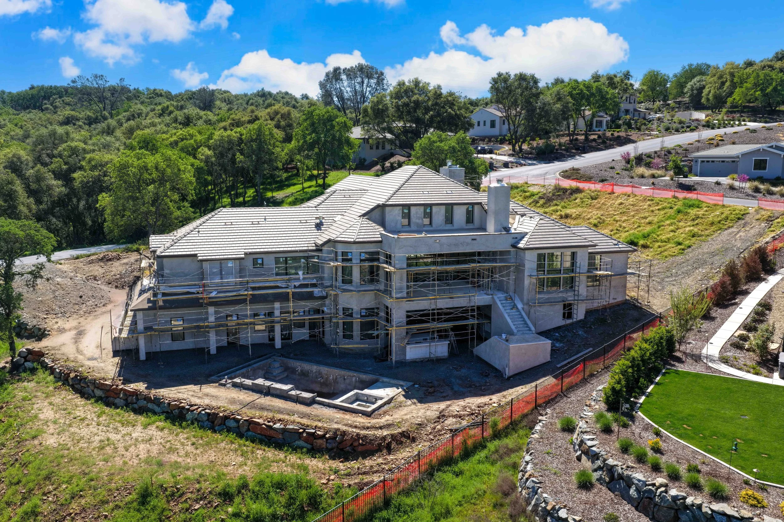 A house under construction on a hillside with visible scaffolding, surrounded by lush green trees and neighboring residential homes, under a bright blue sky.