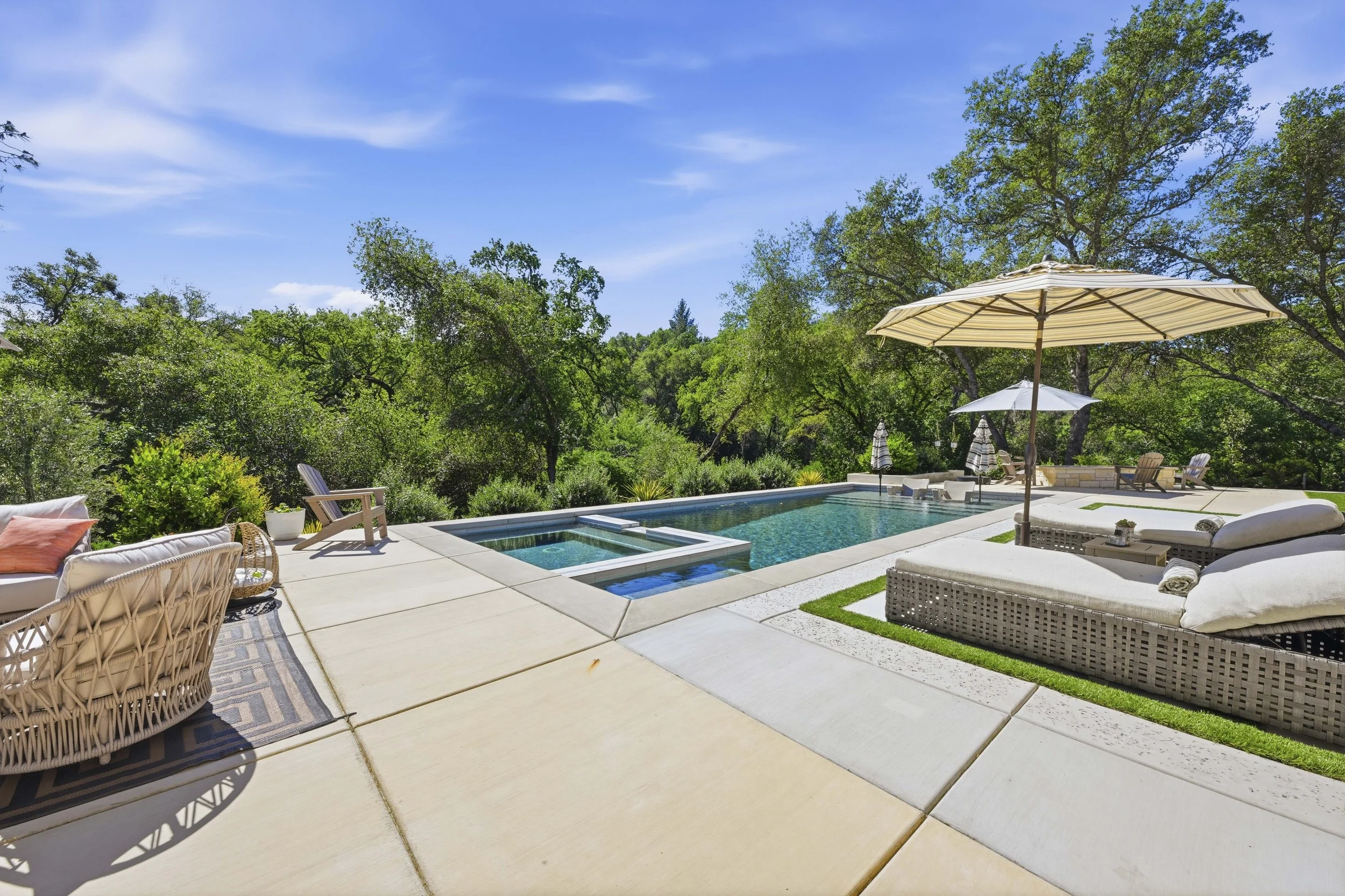 A backyard with a swimming pool, surrounded by lounge chairs, umbrellas, and lush green trees under a blue sky with some clouds.