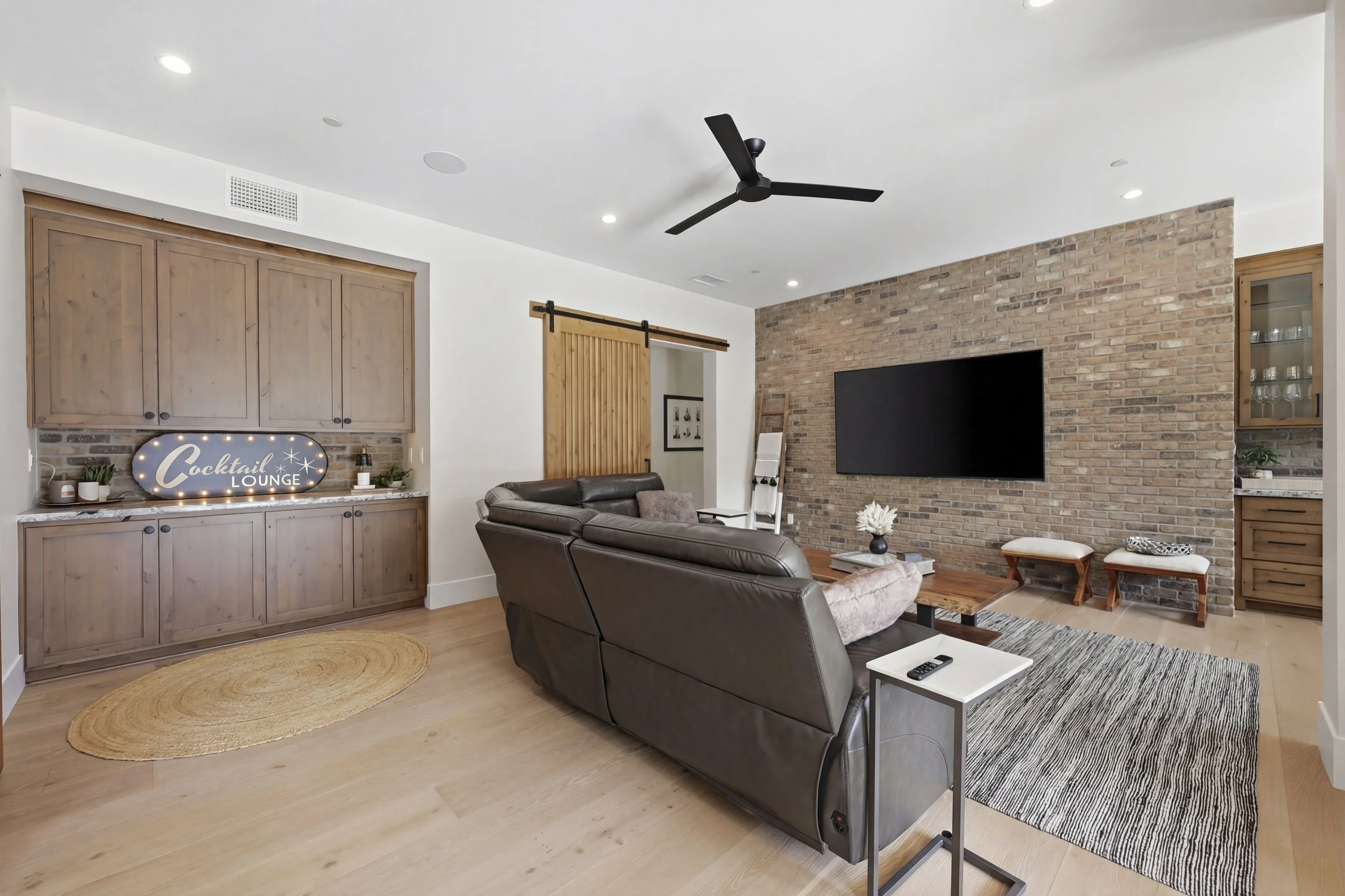 Living room with brick accent wall, mounted large flat-screen TV, leather sofas, ceiling fan, wood cabinets, and decorative items.
