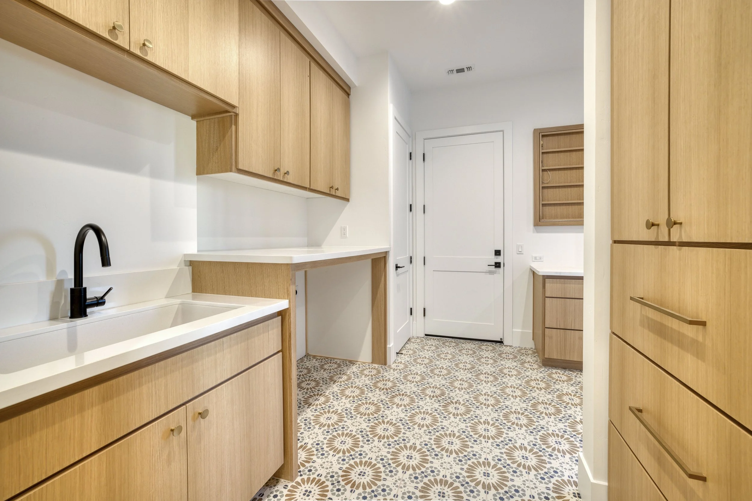 Interior of a modern kitchen with wooden cabinets, a patterned tile floor, a white countertop, and a black faucet at a sink, leading to a white door.