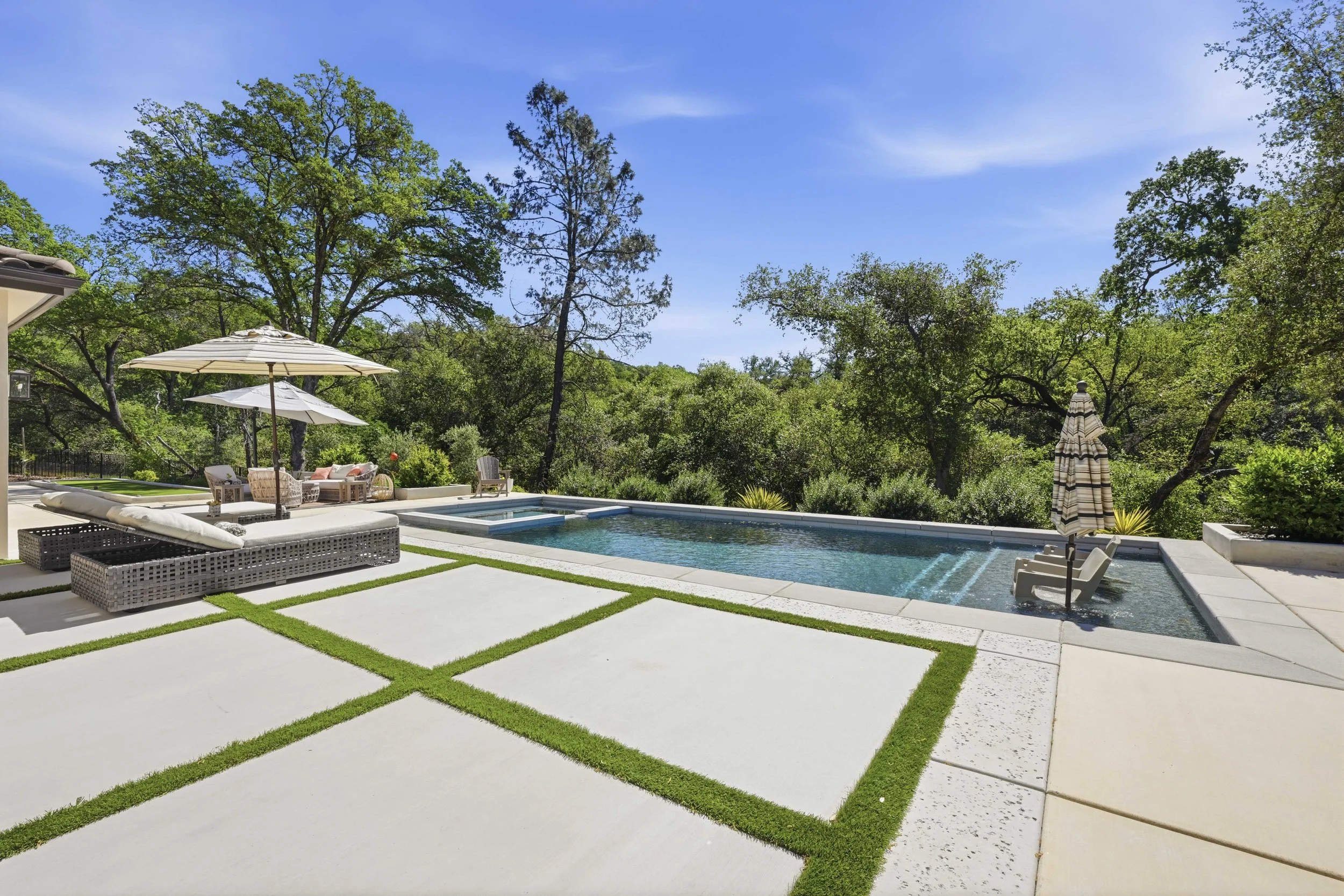 Private backyard with a swimming pool, outdoor seating area with umbrellas, and lush green trees under a blue sky.