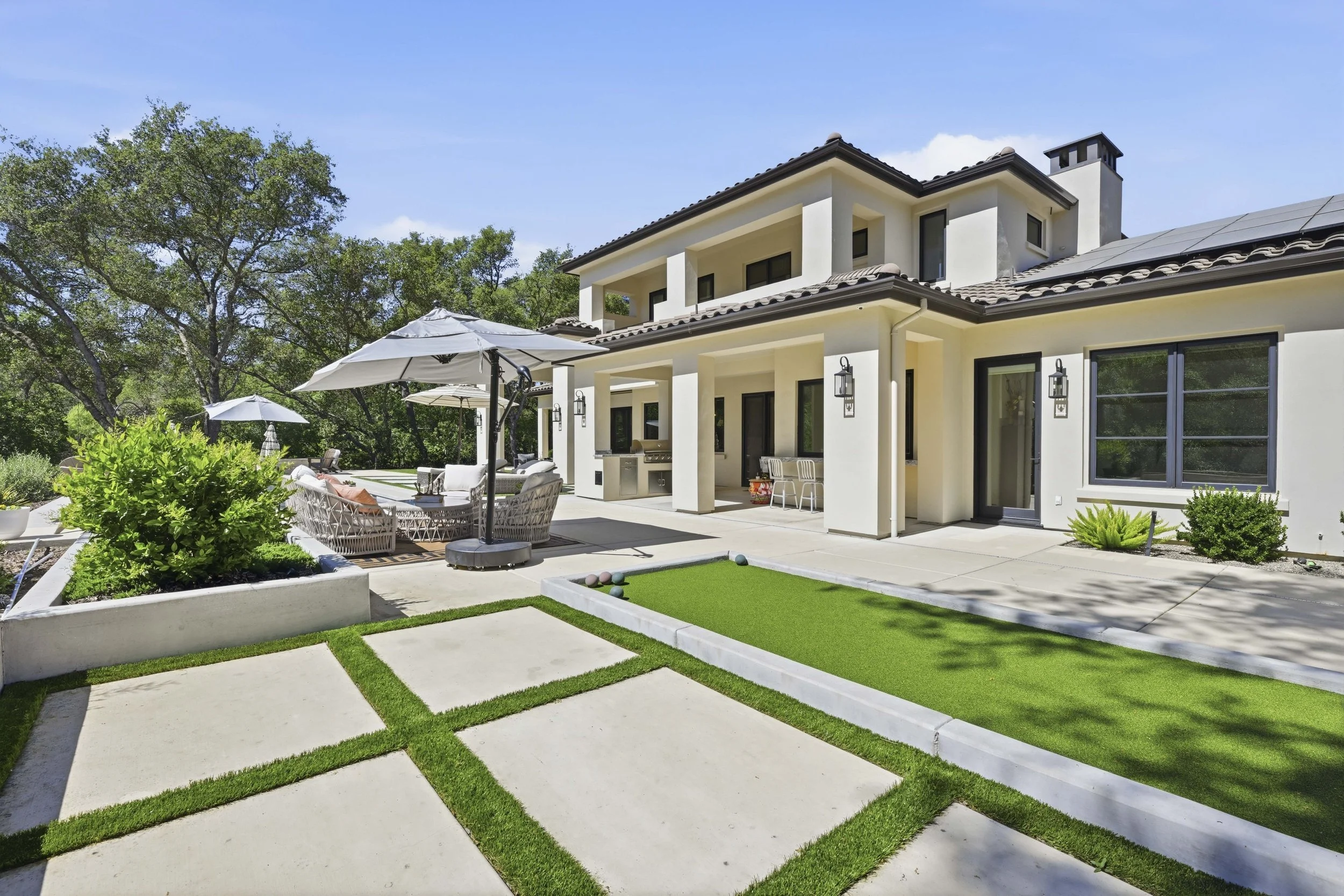 Backyard patio area with outdoor furniture, large umbrellas, a built-in grill, and a small putting green, surrounded by lush greenery and trees.
