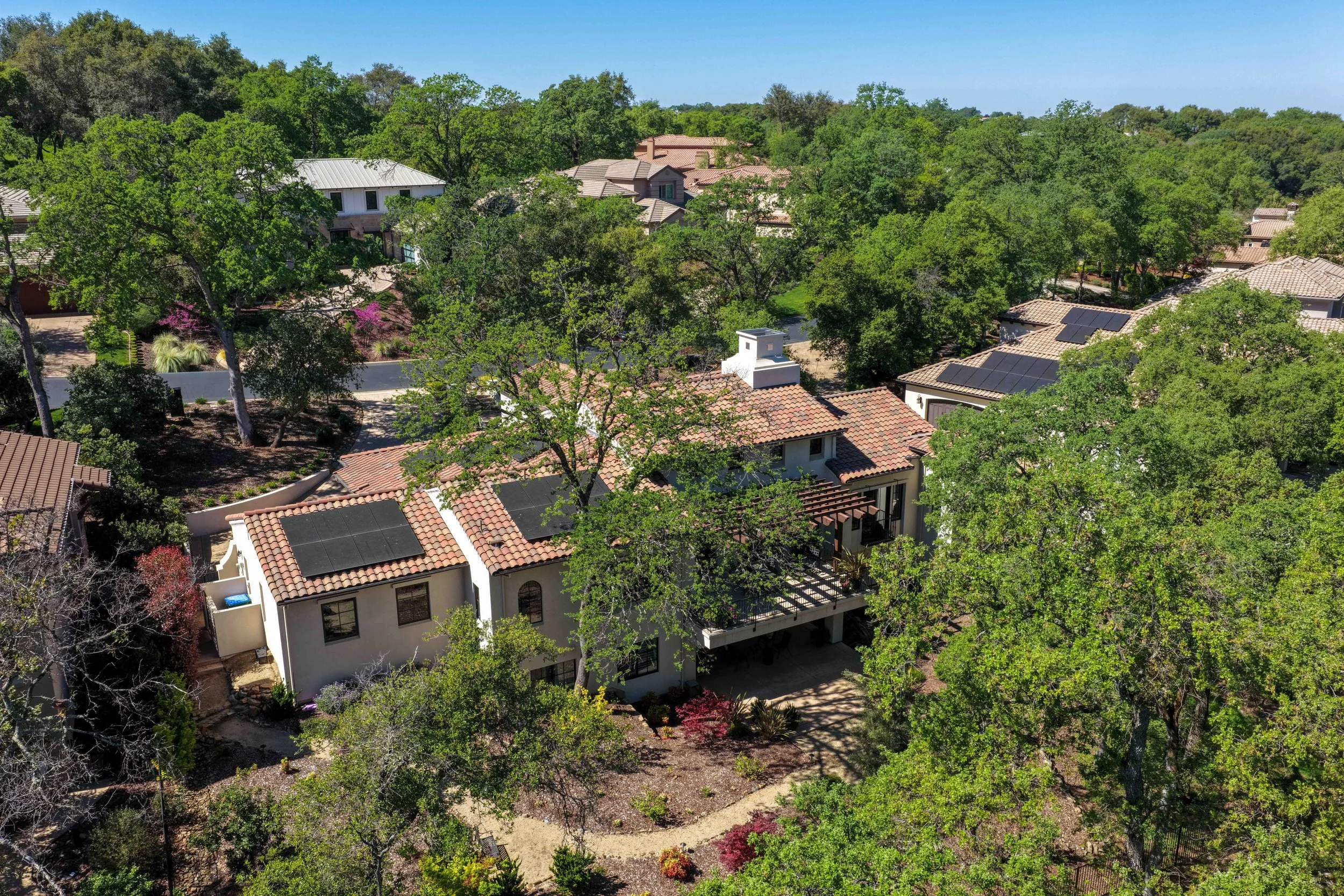 Aerial view of a suburban neighborhood with houses, trees, and solar panels on rooftops under a clear blue sky.