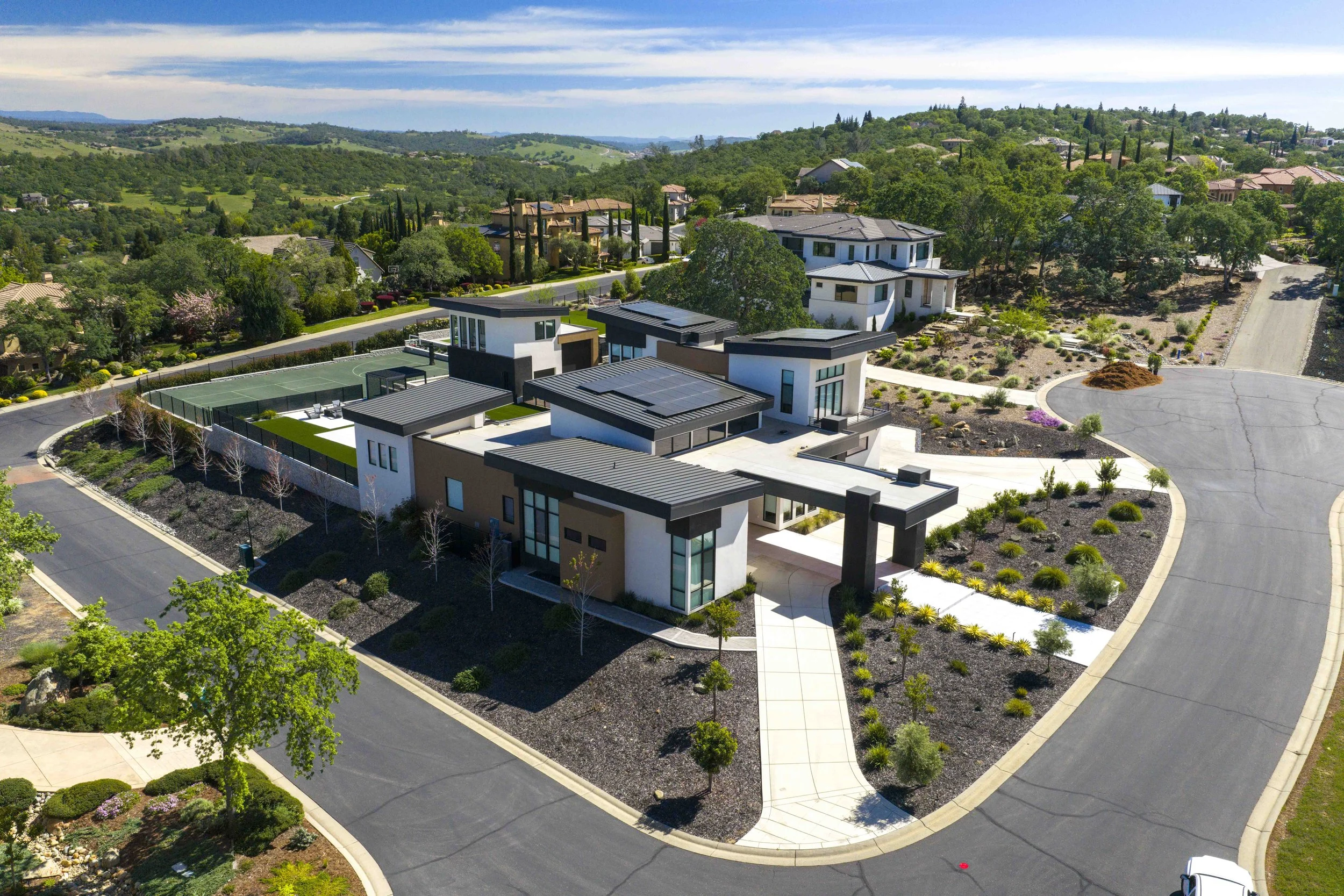 Aerial view of a modern residential neighborhood with contemporary houses, landscaped yards, and a tennis court, set amidst green hills under a partly cloudy sky.